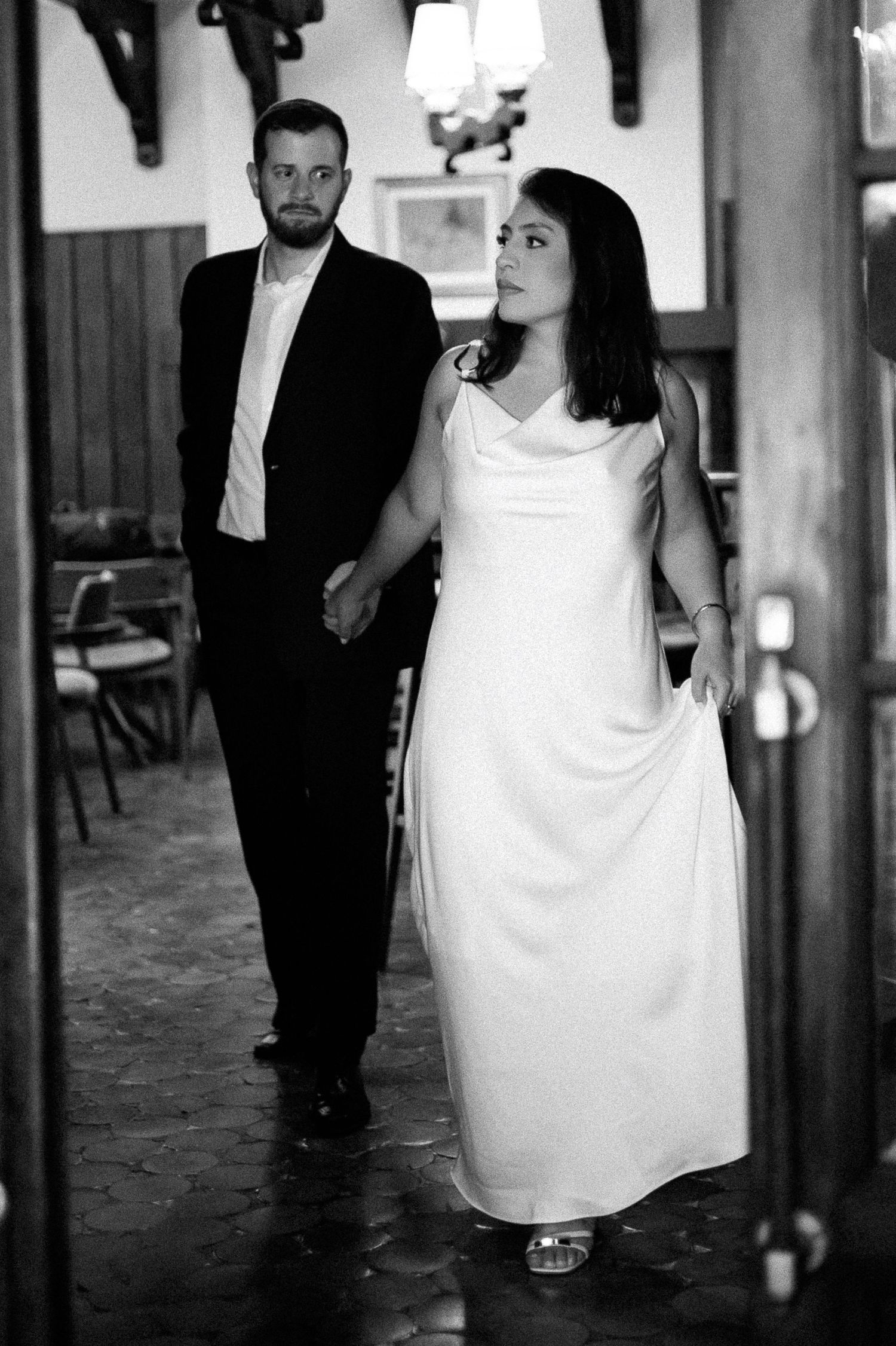 Black and white photo of a bride in white gown and groom in black suit holding hands walking into a venue.