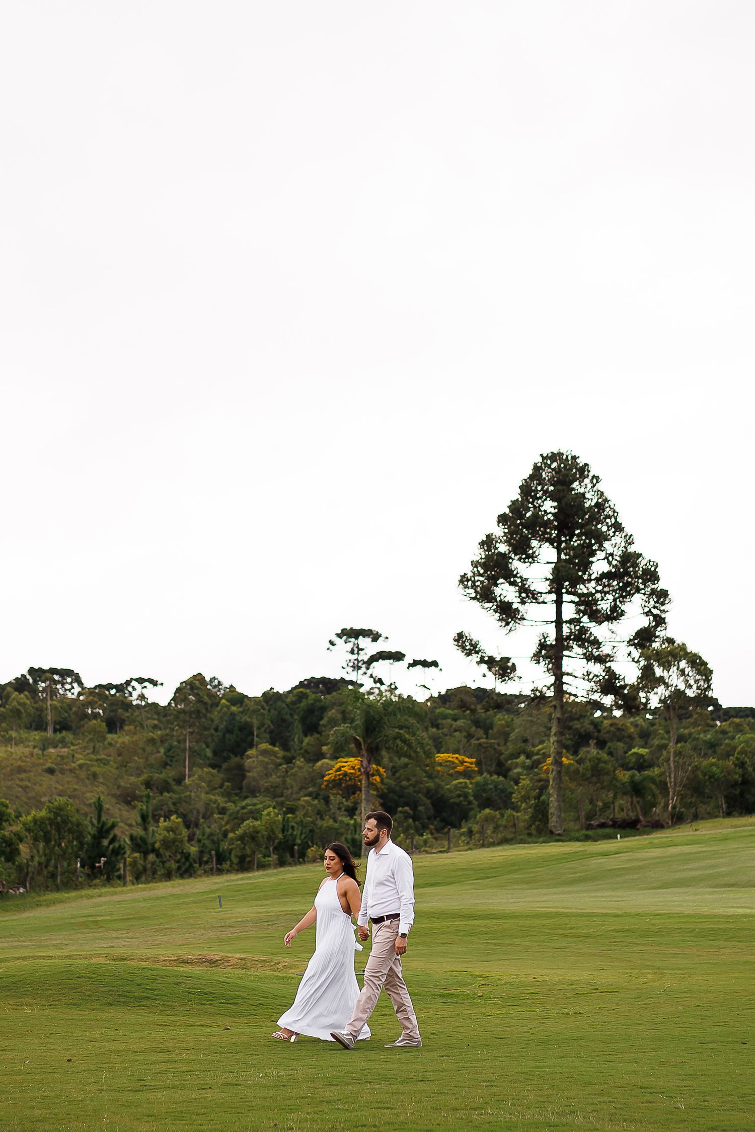 Couple in white outfits walking on lush green golf course with tall pine tree and forest backdrop.