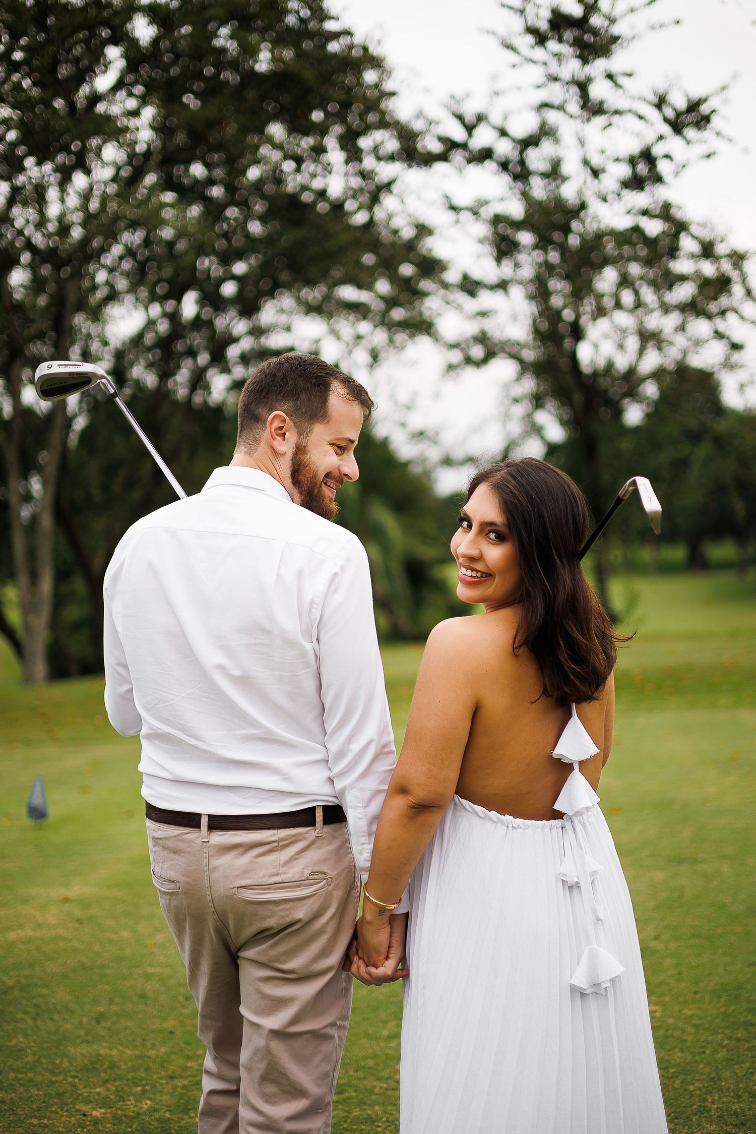 Couple holding hands on golf course, woman in backless white dress smiling over shoulder, man in white shirt with golf clubs.