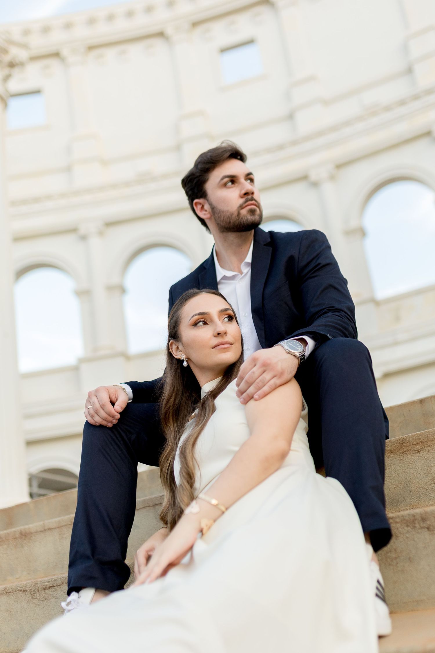 Elegant couple in formal attire sitting on steps near a classical building, gazing upward in a romantic pose.