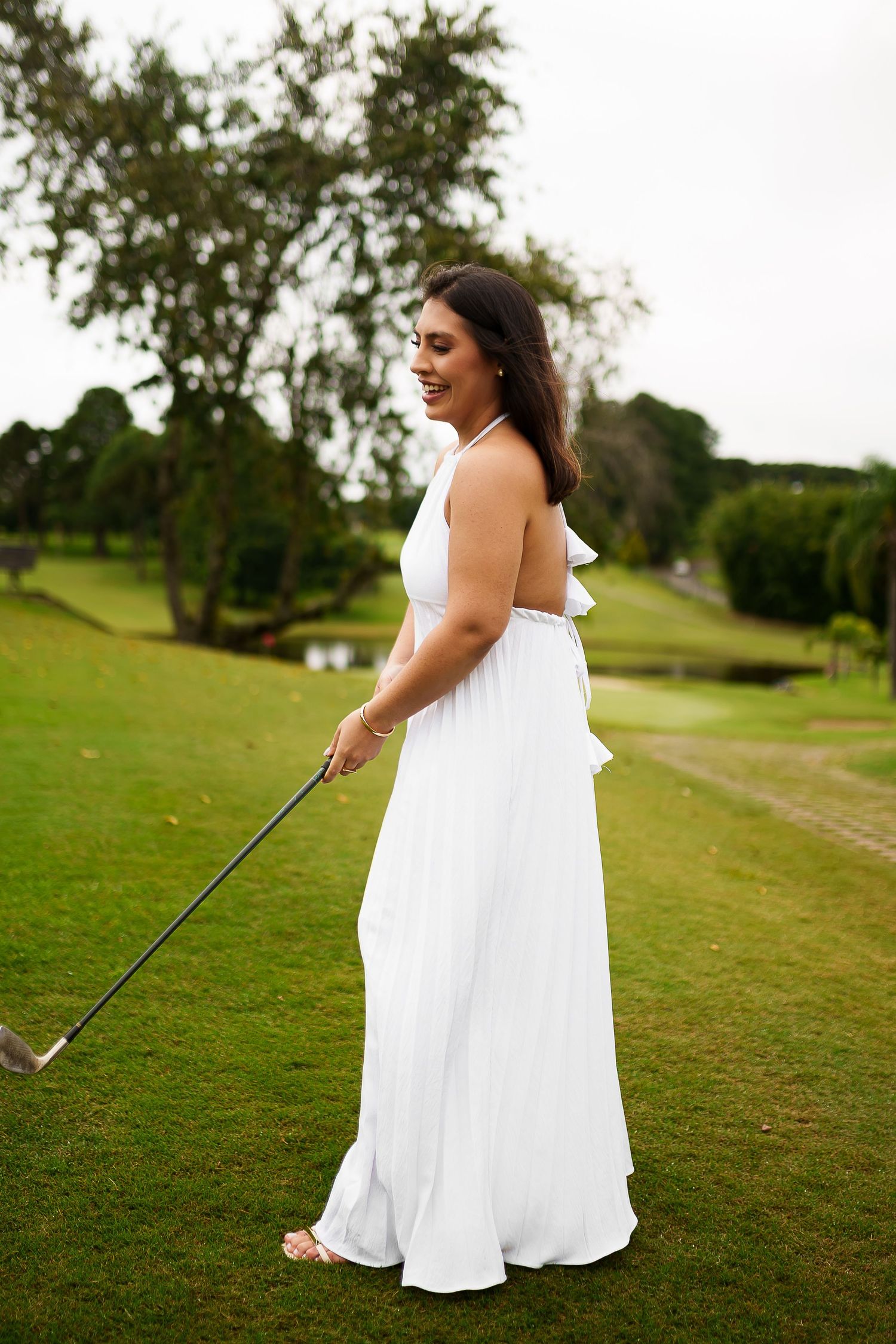 Woman in white halter maxi dress holding a golf club on a lush green golf course.