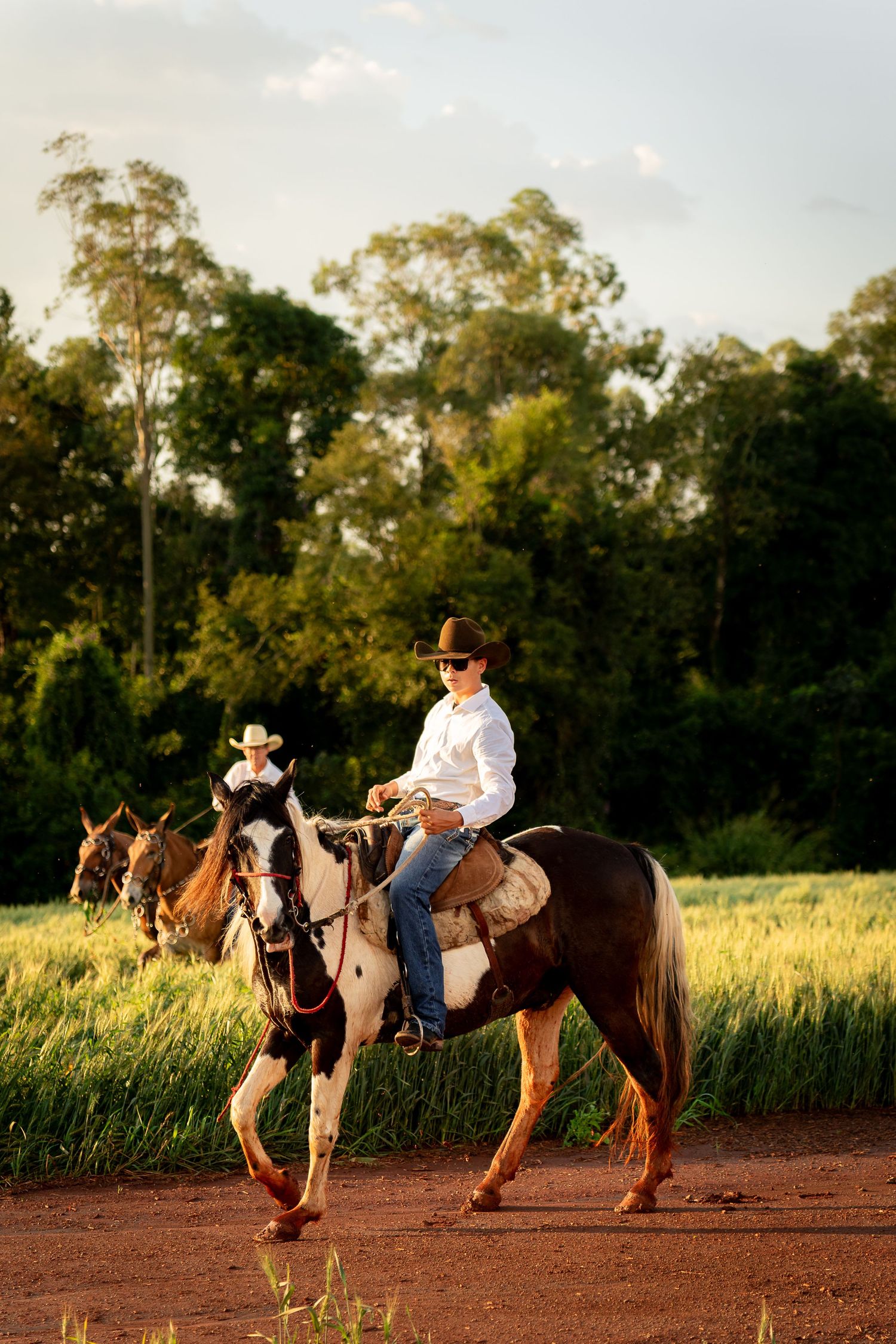 Two cowboys in hats ride horses along a dirt path beside green fields at golden hour.