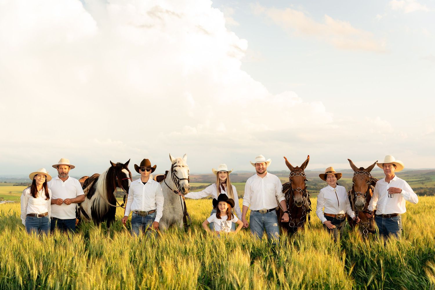 A family in cowboy hats poses with horses and mules in a lush green field under a dramatic cloudy sky.