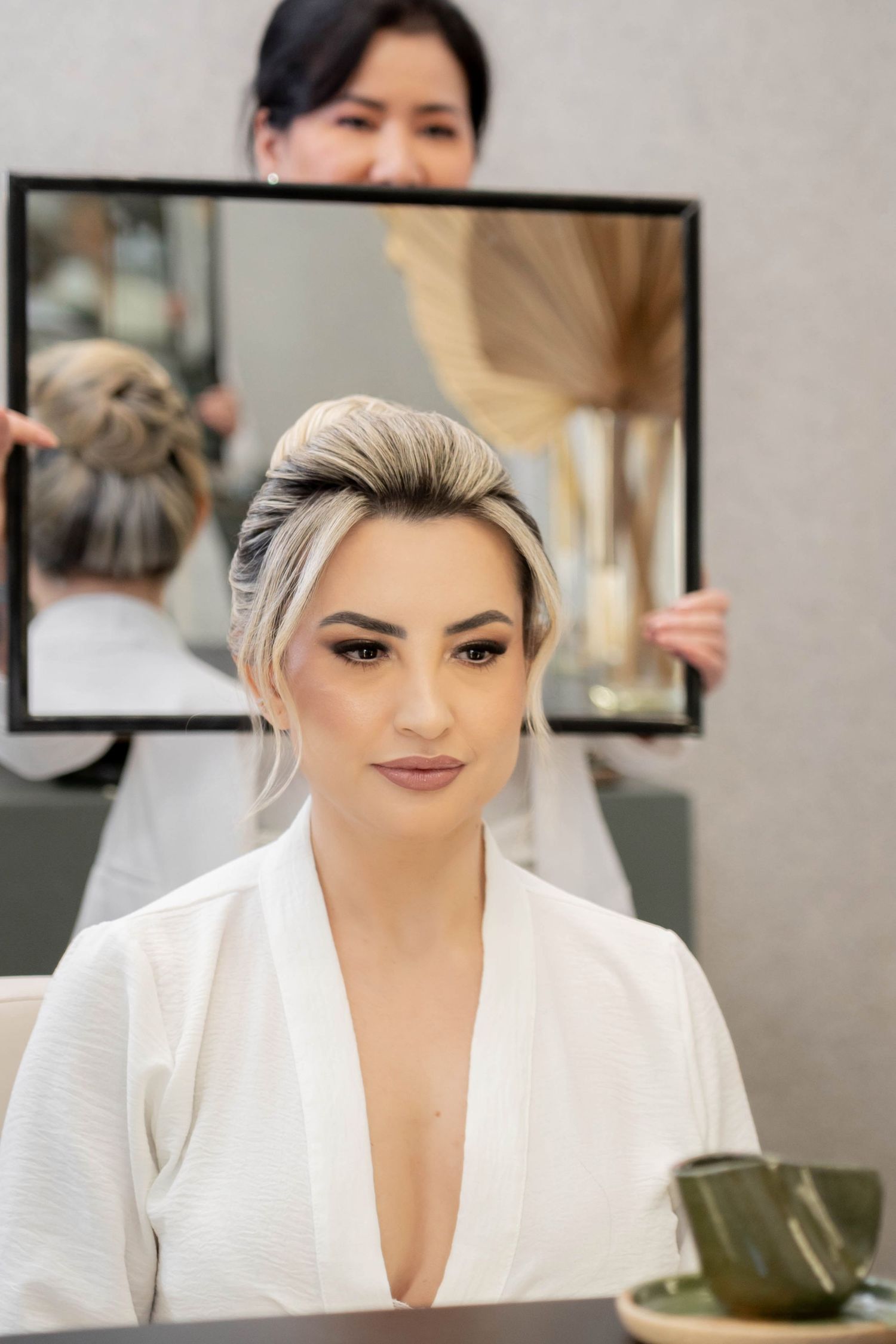 Bride in white robe with elegant updo hairstyle gets final look revealed in mirror at bridal salon.