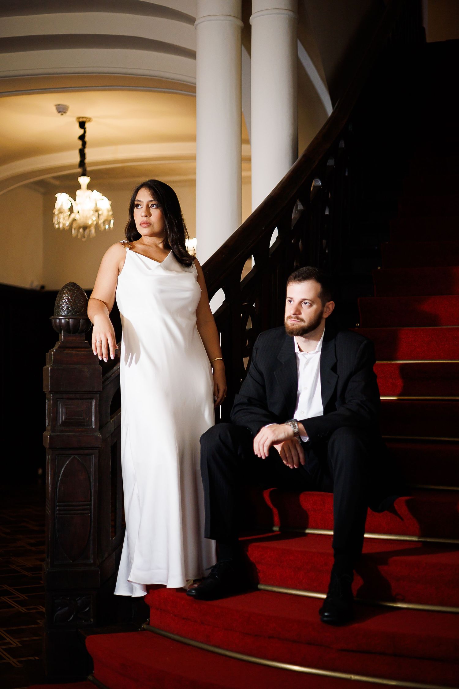 A woman in a white gown and a man in a black suit pose elegantly on red-carpeted stairs in a grand interior.