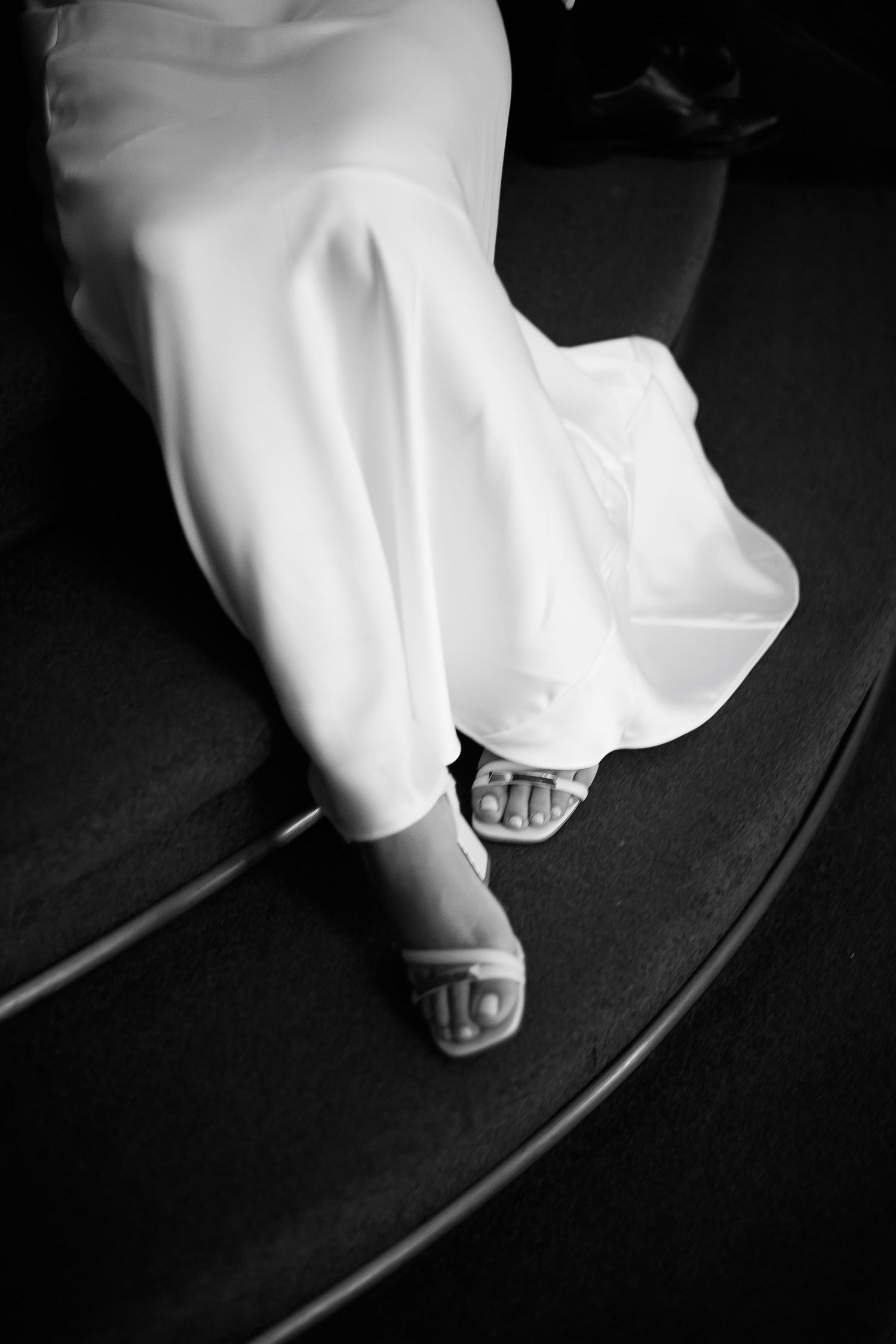 Black and white photo of a bride in a white gown wearing elegant sandal heels, seated on dark steps.