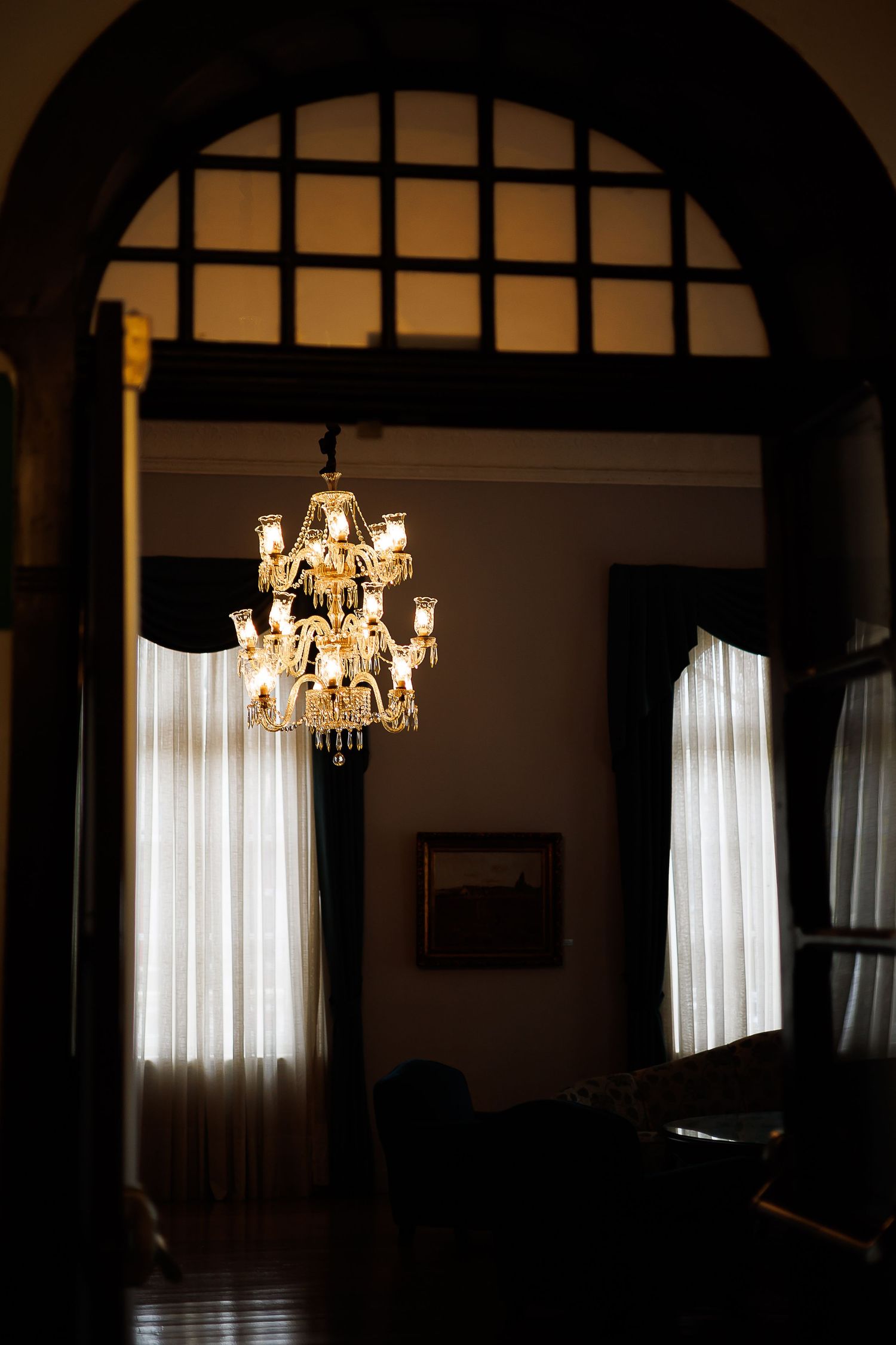 Elegant crystal chandelier hanging in a dimly lit vintage room with arched windows and sheer white curtains.