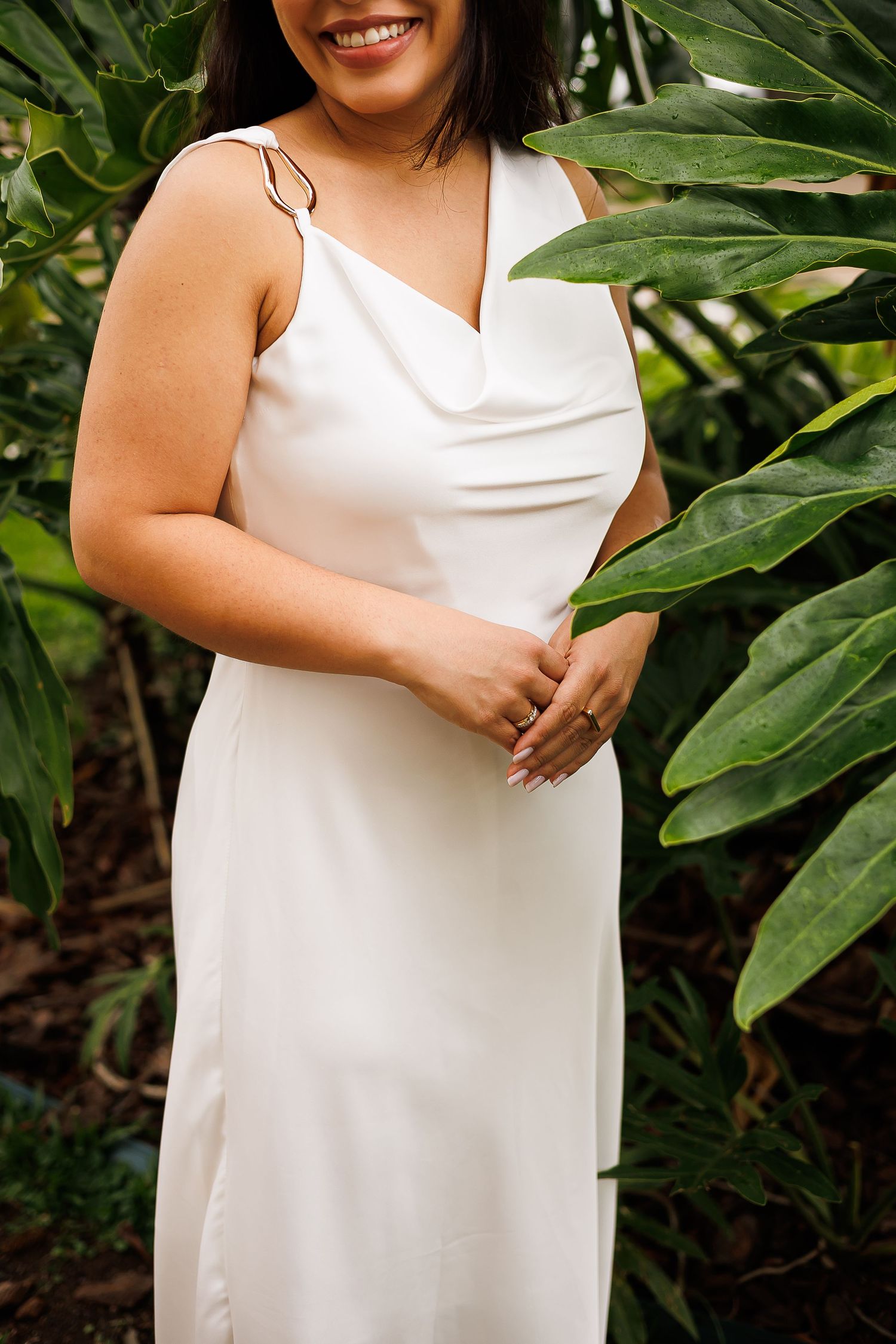Smiling bride in a white cowl neck satin slip wedding dress standing among tropical green leaves.