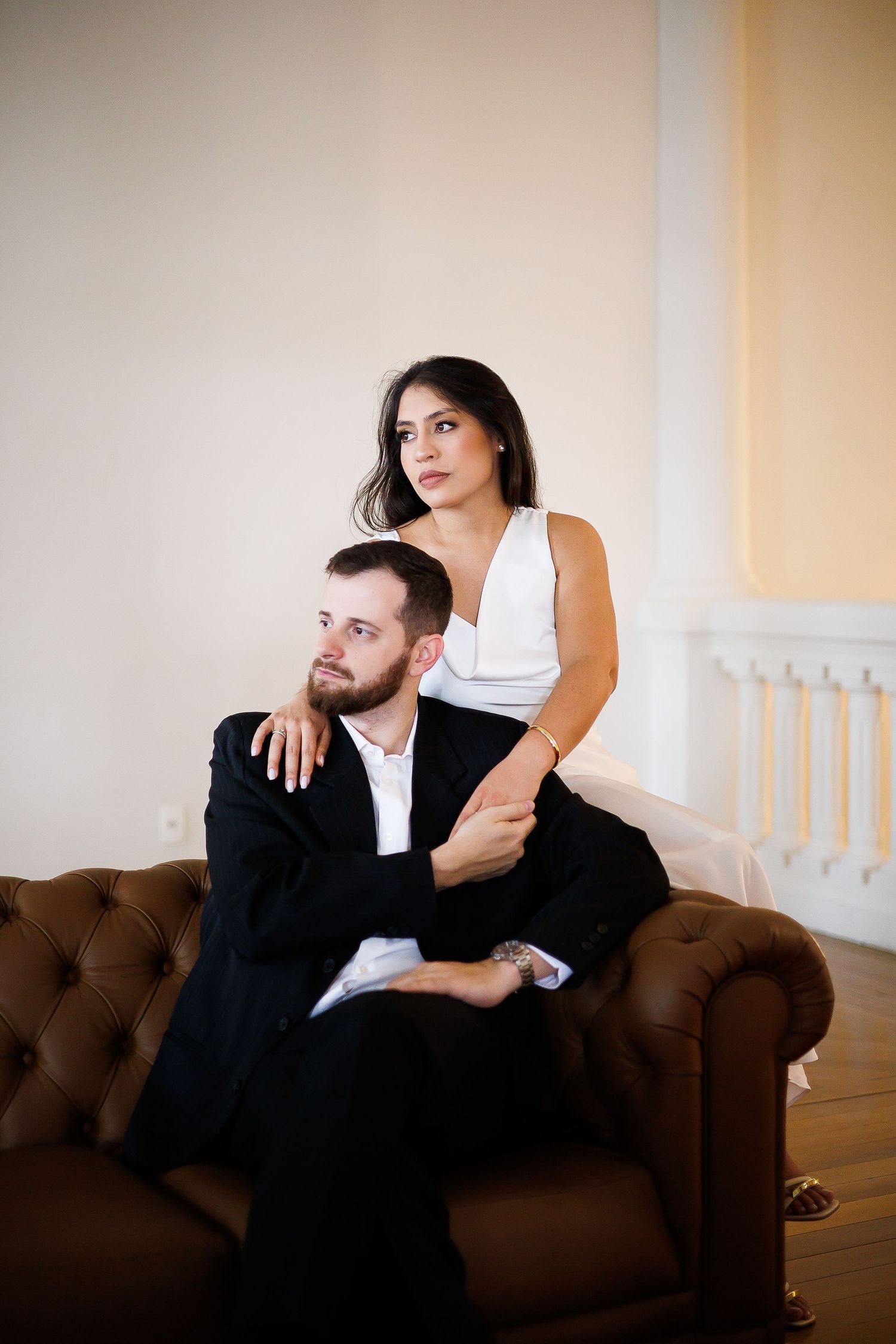 Elegant couple posing on a brown leather sofa, man in black suit seated while woman in white dress stands behind him.