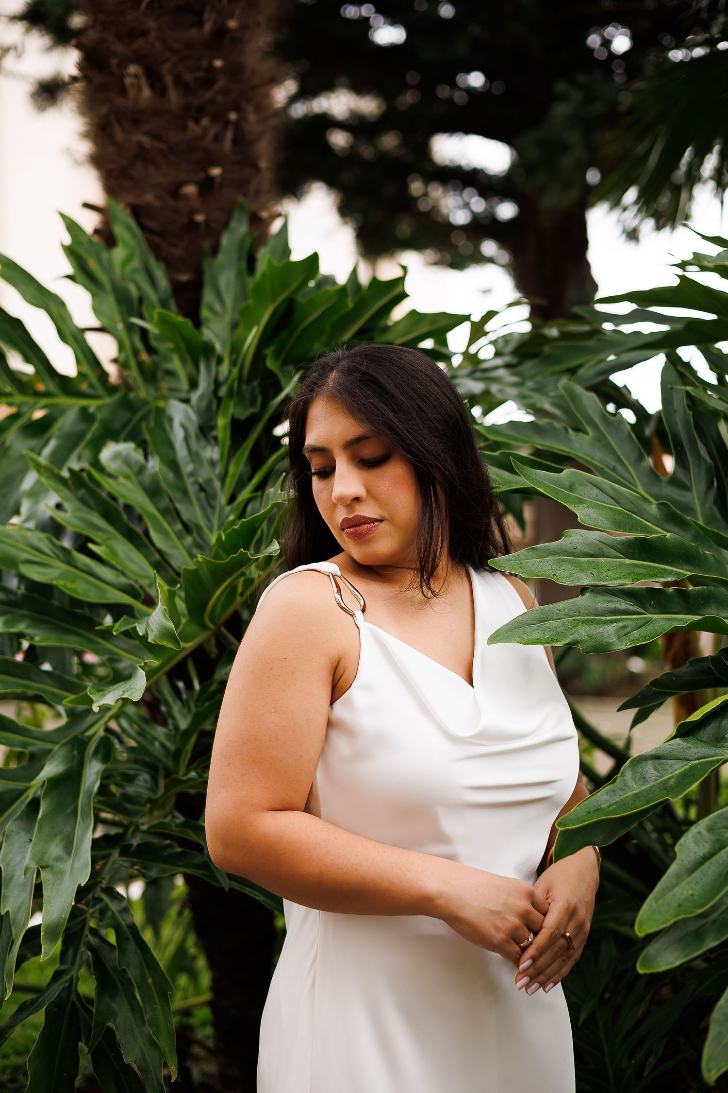 Woman in white satin slip dress poses among lush tropical green leaves and palm trees in an elegant outdoor setting.