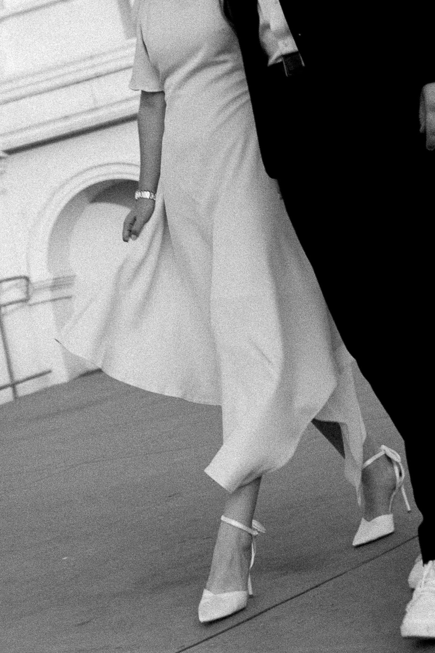 Black and white photo of a woman in a flowing white dress and strappy heels walking gracefully on a runway.