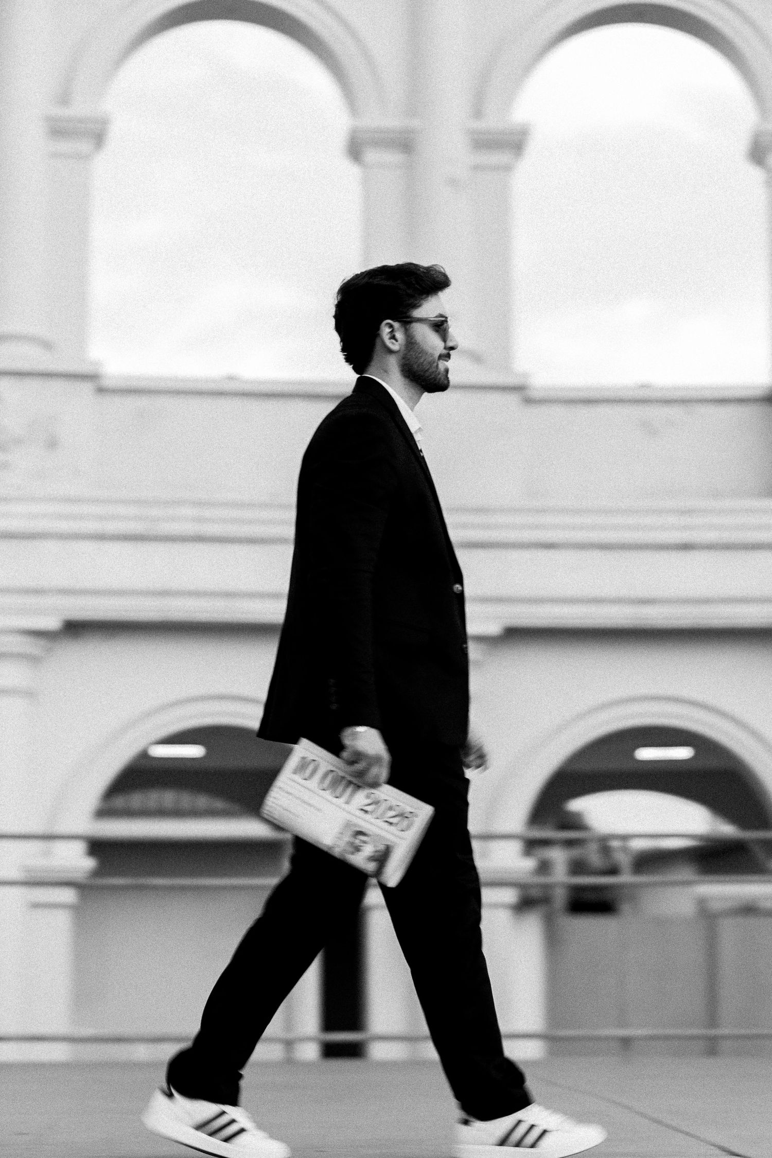 A stylish man in a black suit and Adidas sneakers walks confidently while holding a newspaper outdoors.