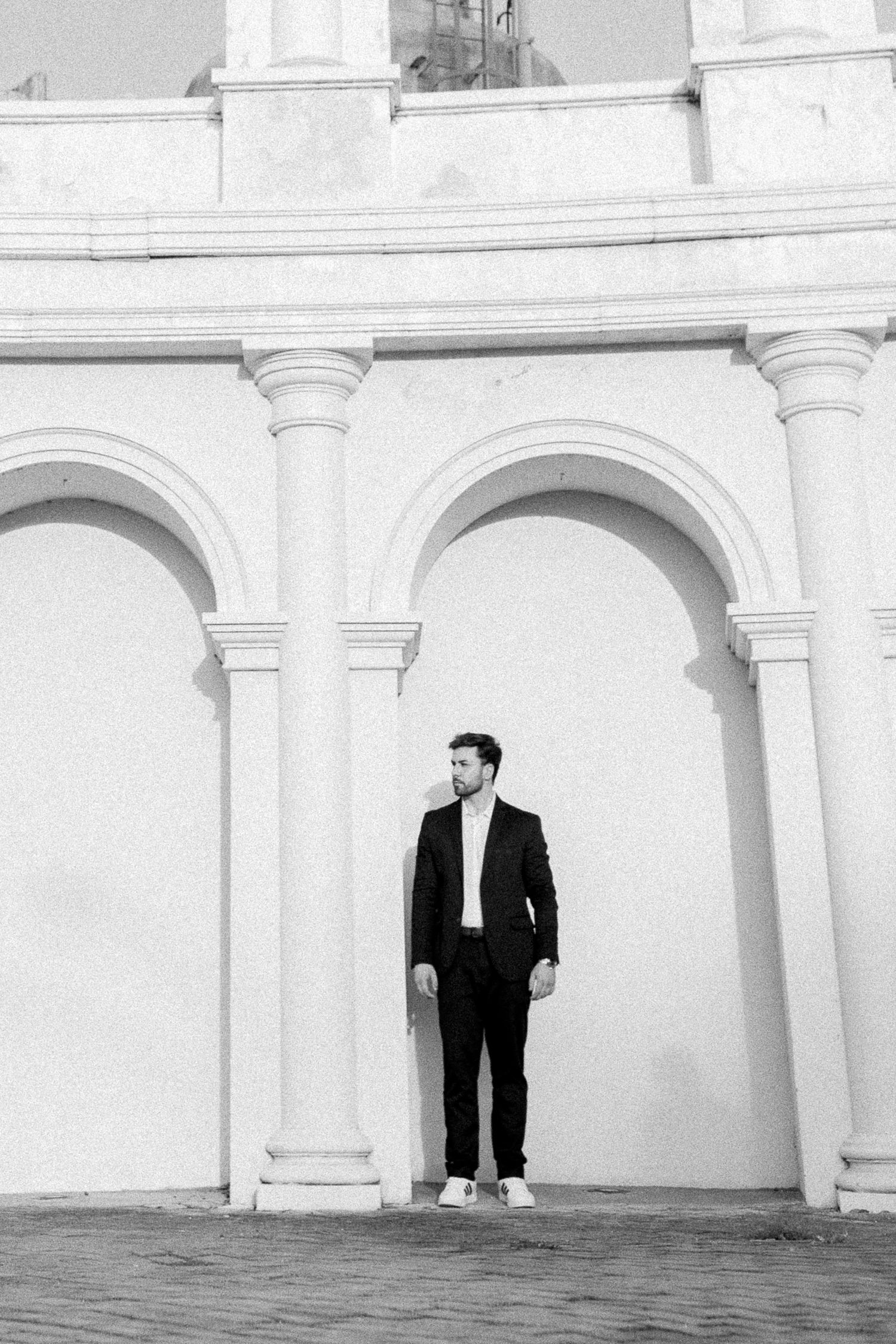 Black and white photo of a bearded man in a dark suit standing beneath elegant arched columns of a classical building.