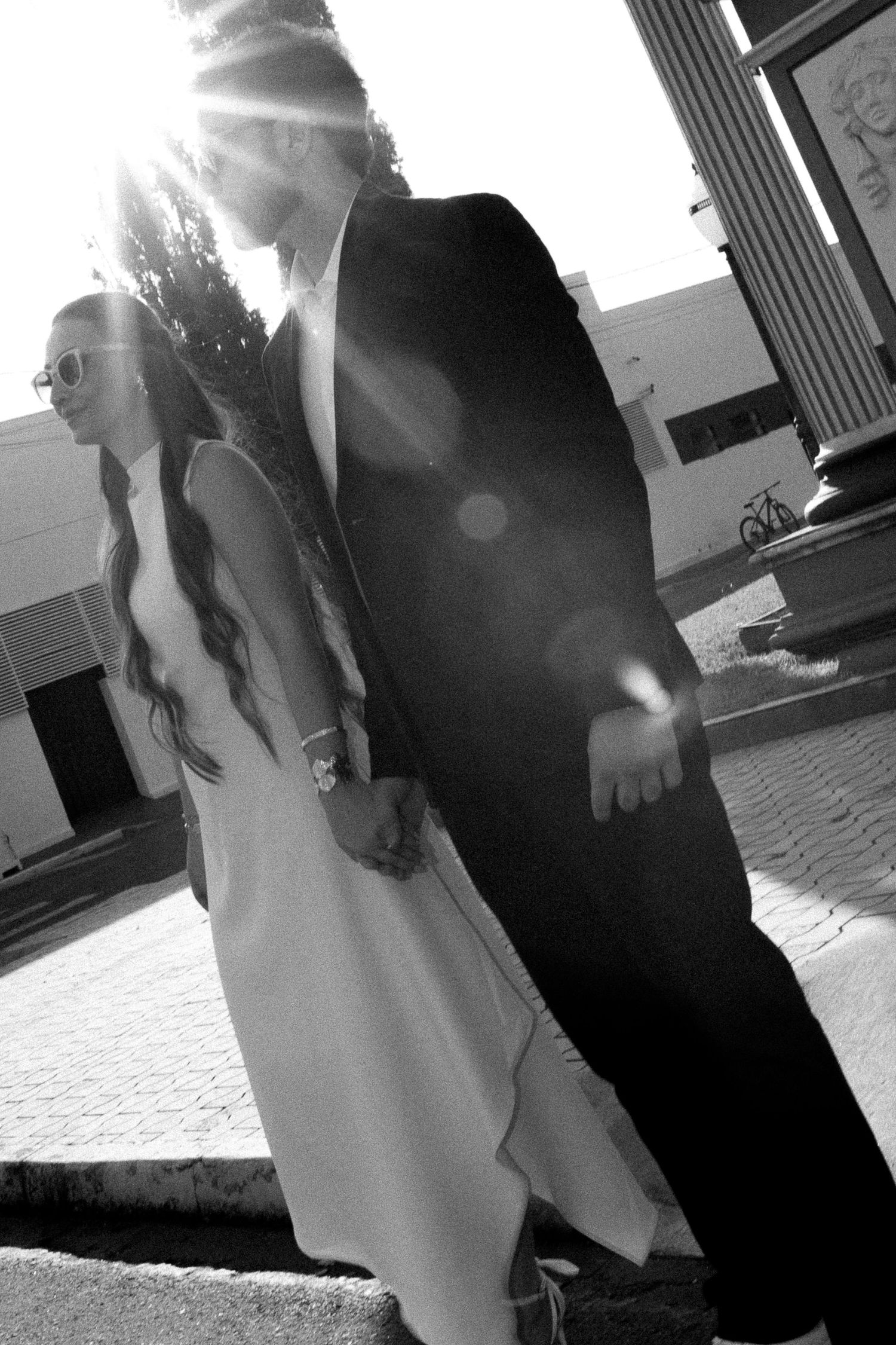 Black and white photo of a couple in wedding attire holding hands outdoors with sun flare behind them.