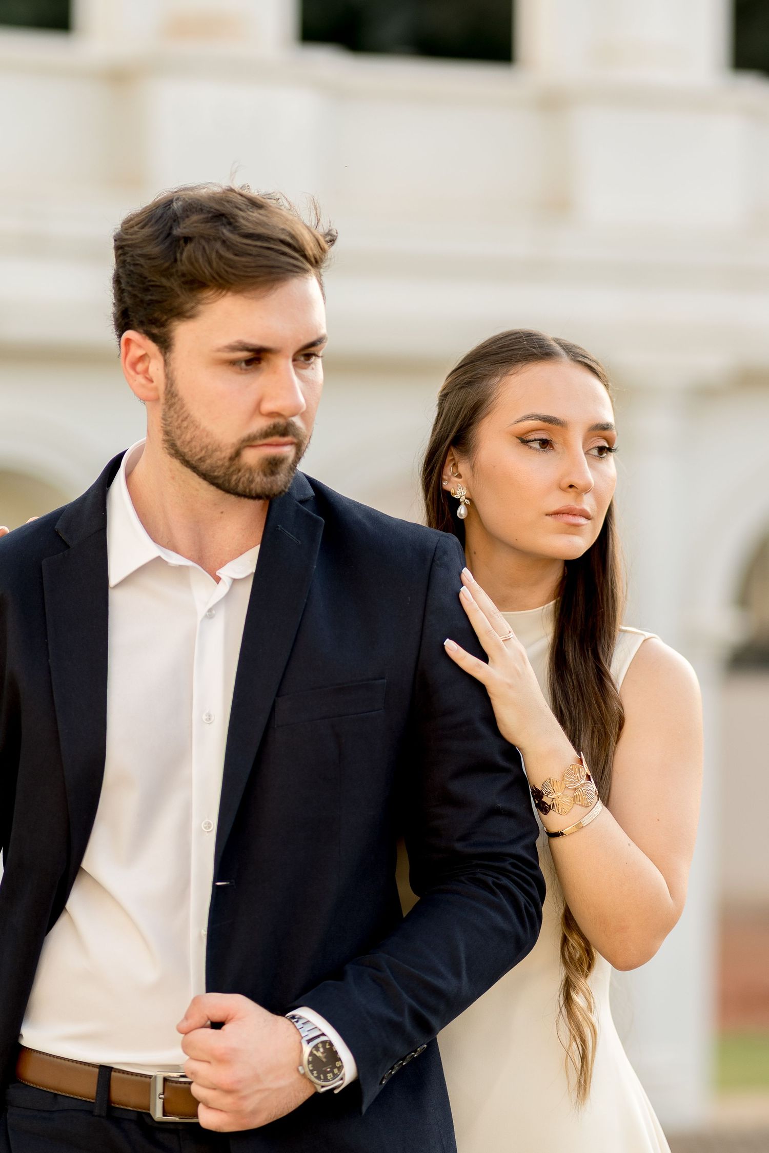 Stylish couple in formal attire walking near classical white building, woman holding mans arm in cream dress.