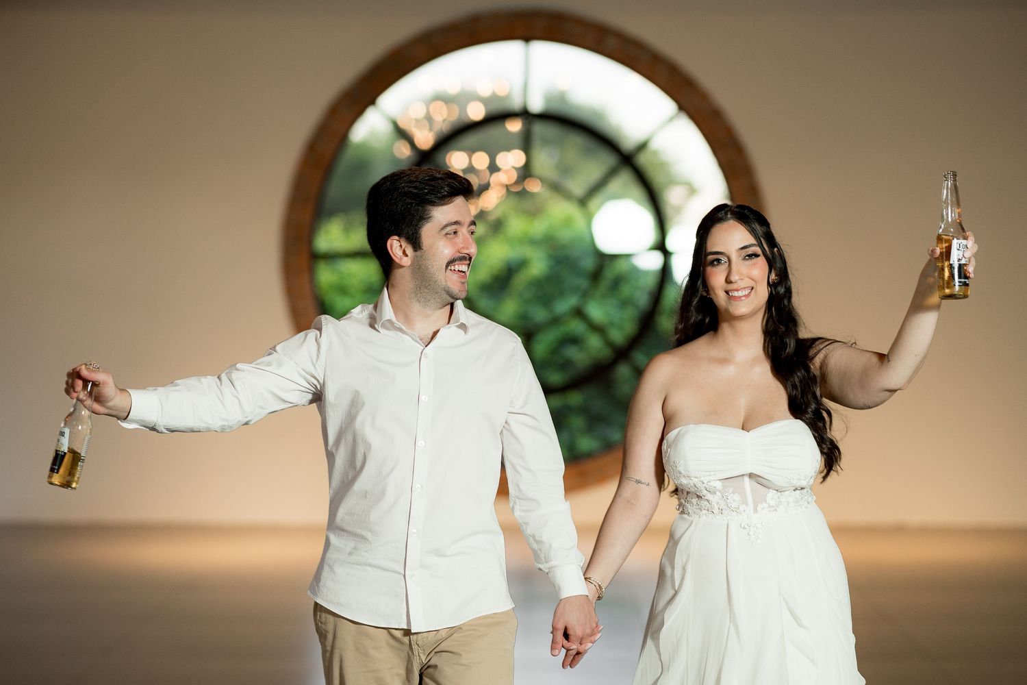 Couple in white attire holding Corona beers, posing in front of a round window at their wedding celebration.