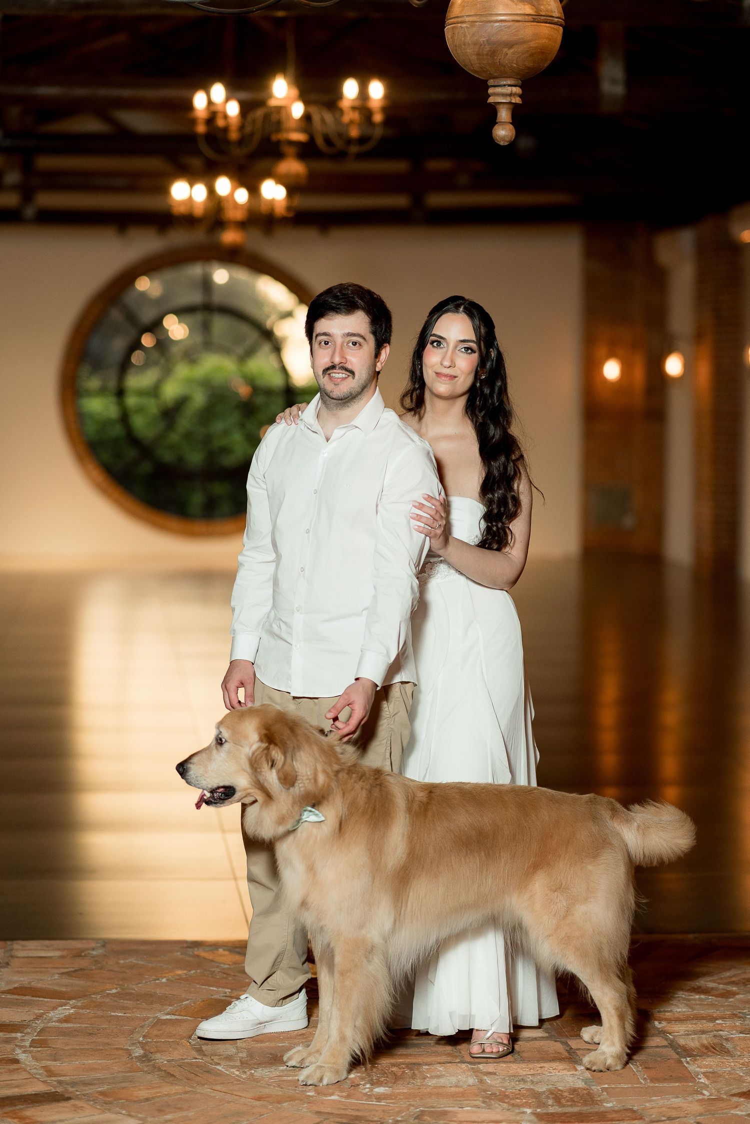 Couple in white attire poses with a golden retriever inside an elegant venue with chandelier lighting.