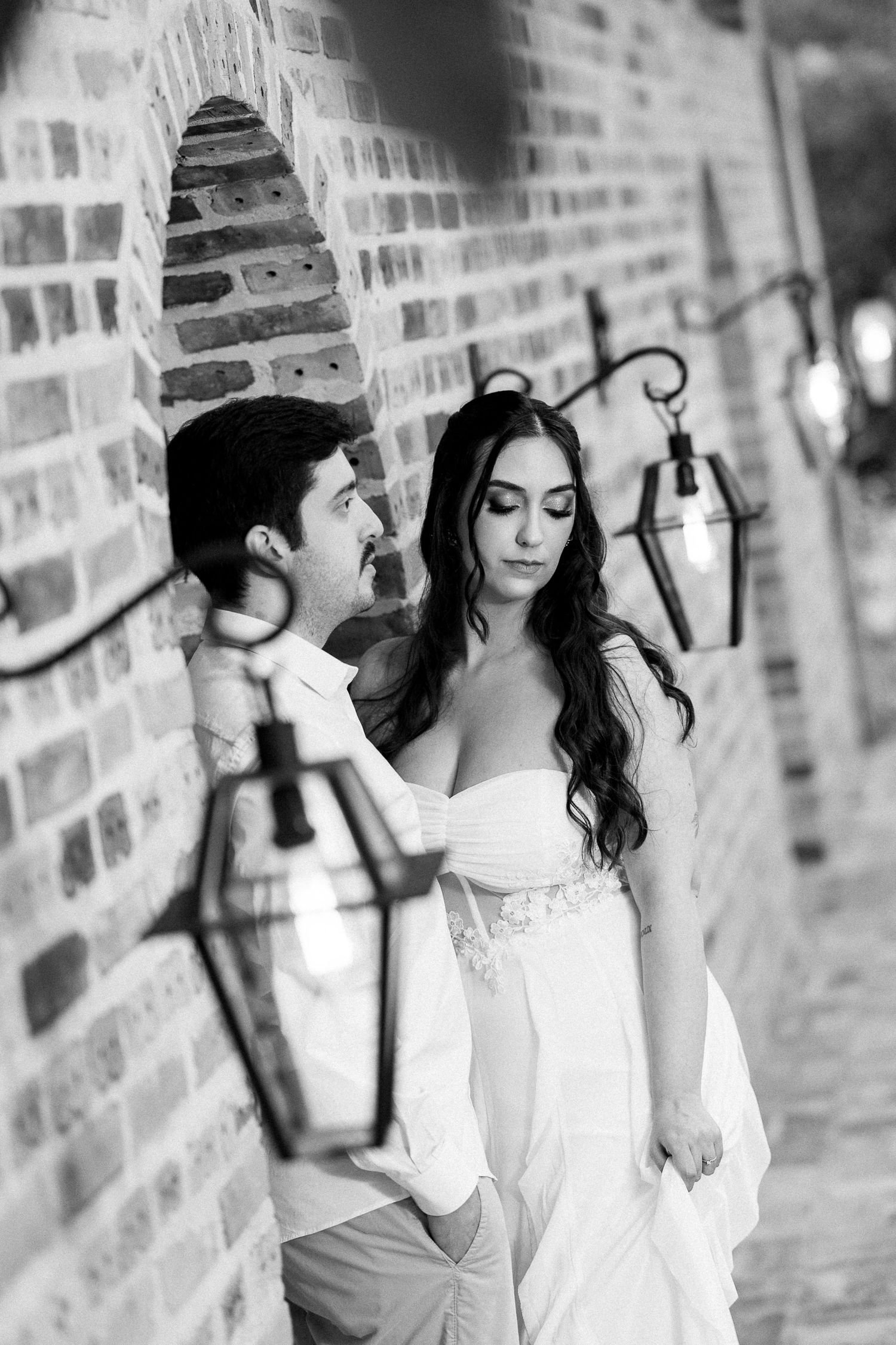 Black and white photo of a bride and groom posing against a brick wall with vintage lanterns at their wedding.