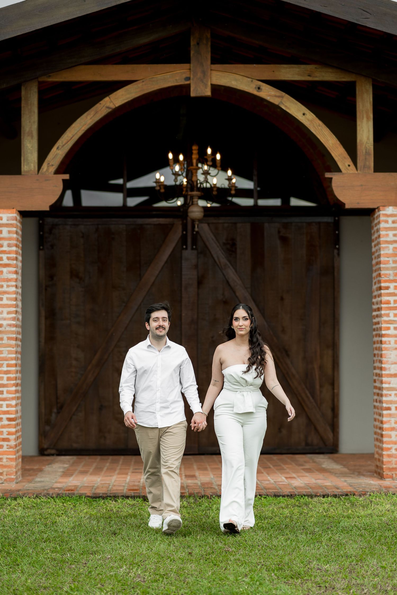 Couple holding hands walking on grass in front of rustic barn doors with wooden arch and chandelier.