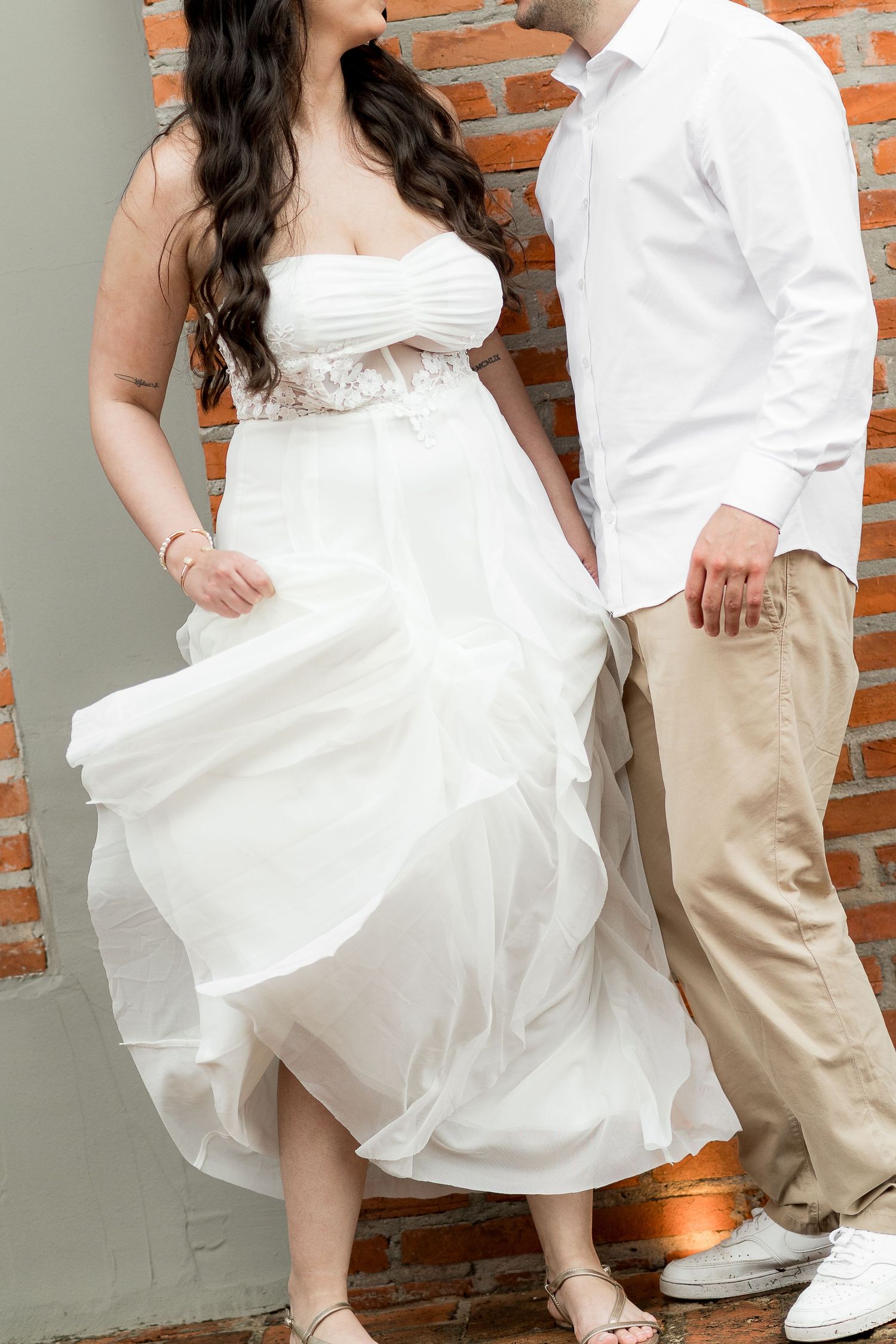 Couple posing against brick wall, bride in flowing white strapless gown, groom in white shirt and khaki pants.