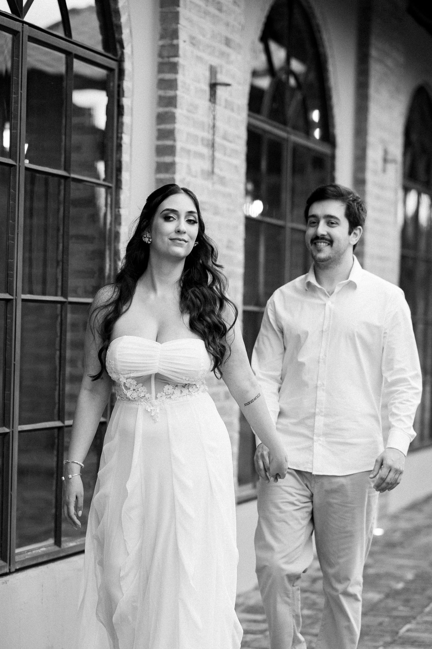 Black and white photo of a smiling couple in wedding attire holding hands while walking near a brick building.