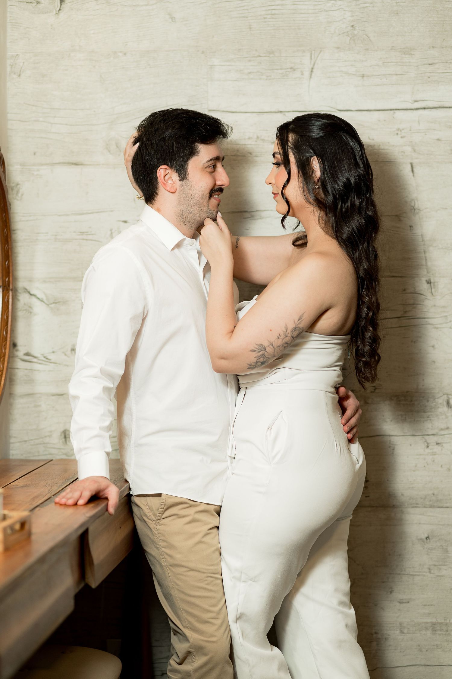 A couple in white outfits share a romantic moment, the woman touching the mans face while smiling at each other.