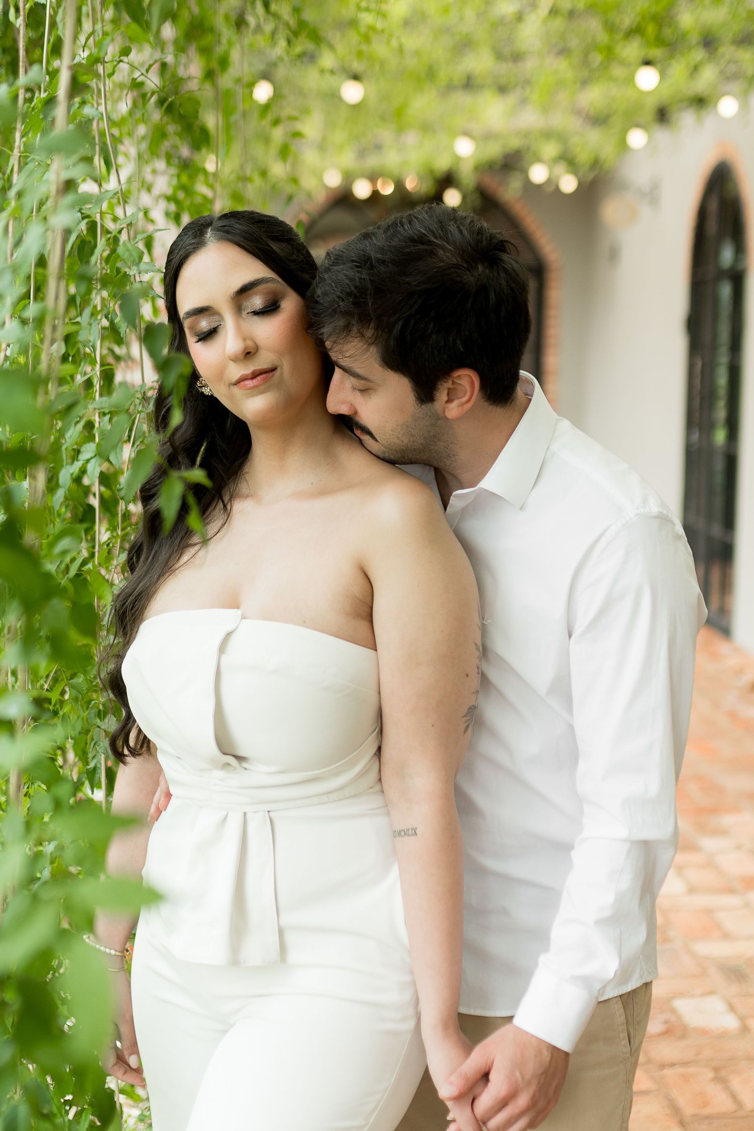 Couple in white attire embracing outdoors under string lights and lush green vines at an elegant venue.