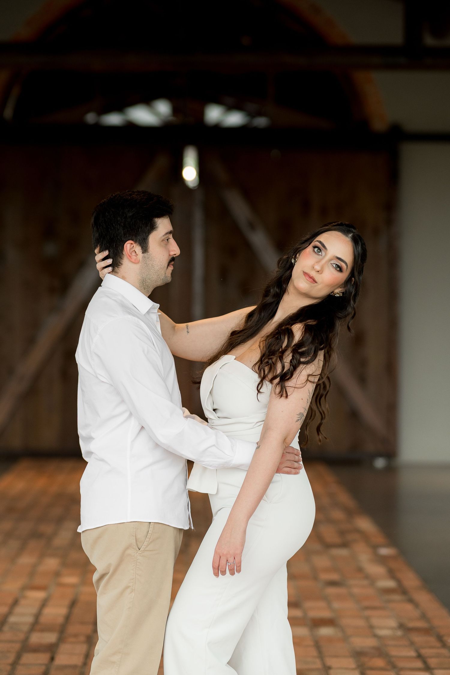 Couple in white attire embrace in a rustic barn venue, bride gazing up at groom during wedding photo session.