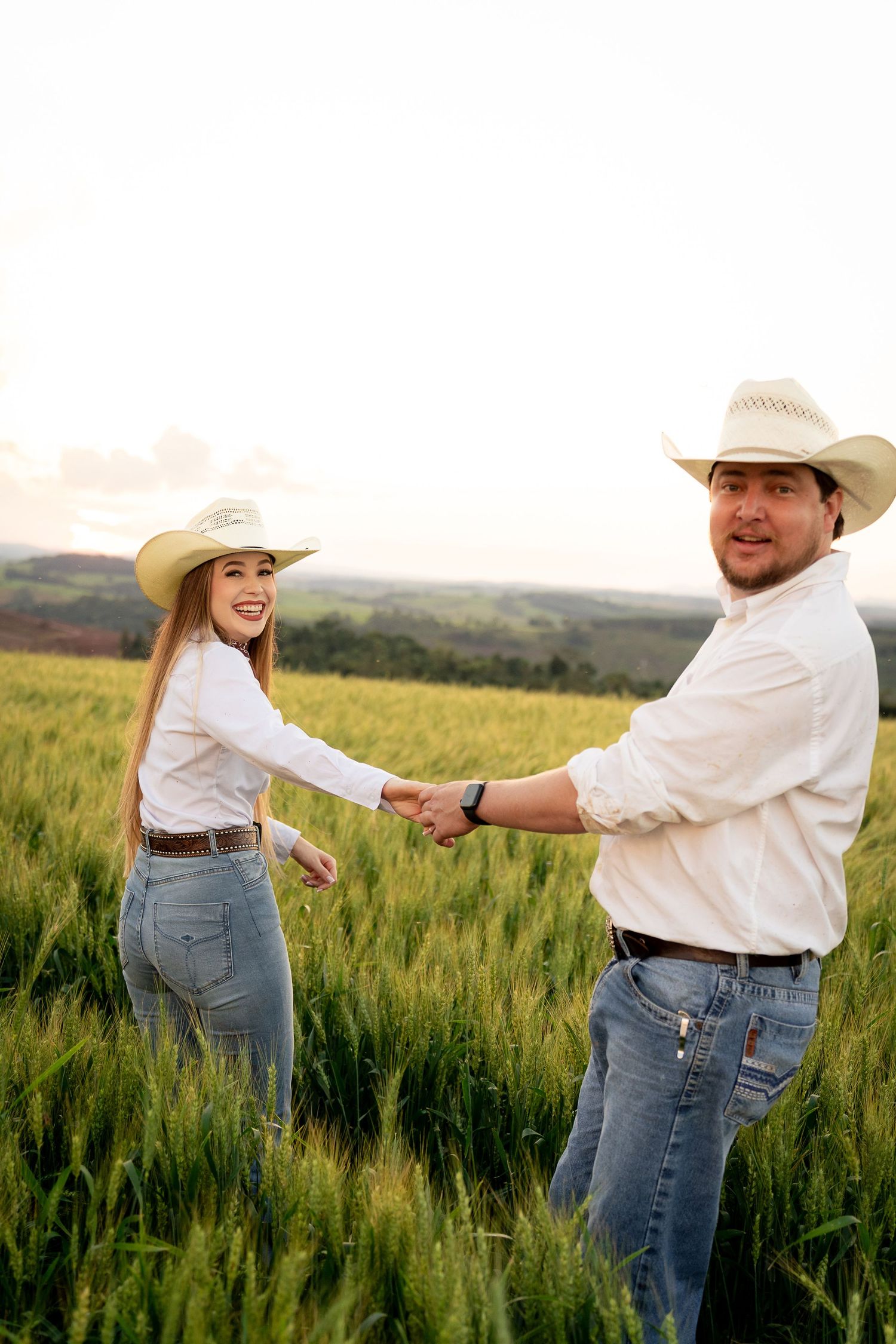 Couple in white shirts and cowboy hats holding hands in a lush green wheat field at sunset.