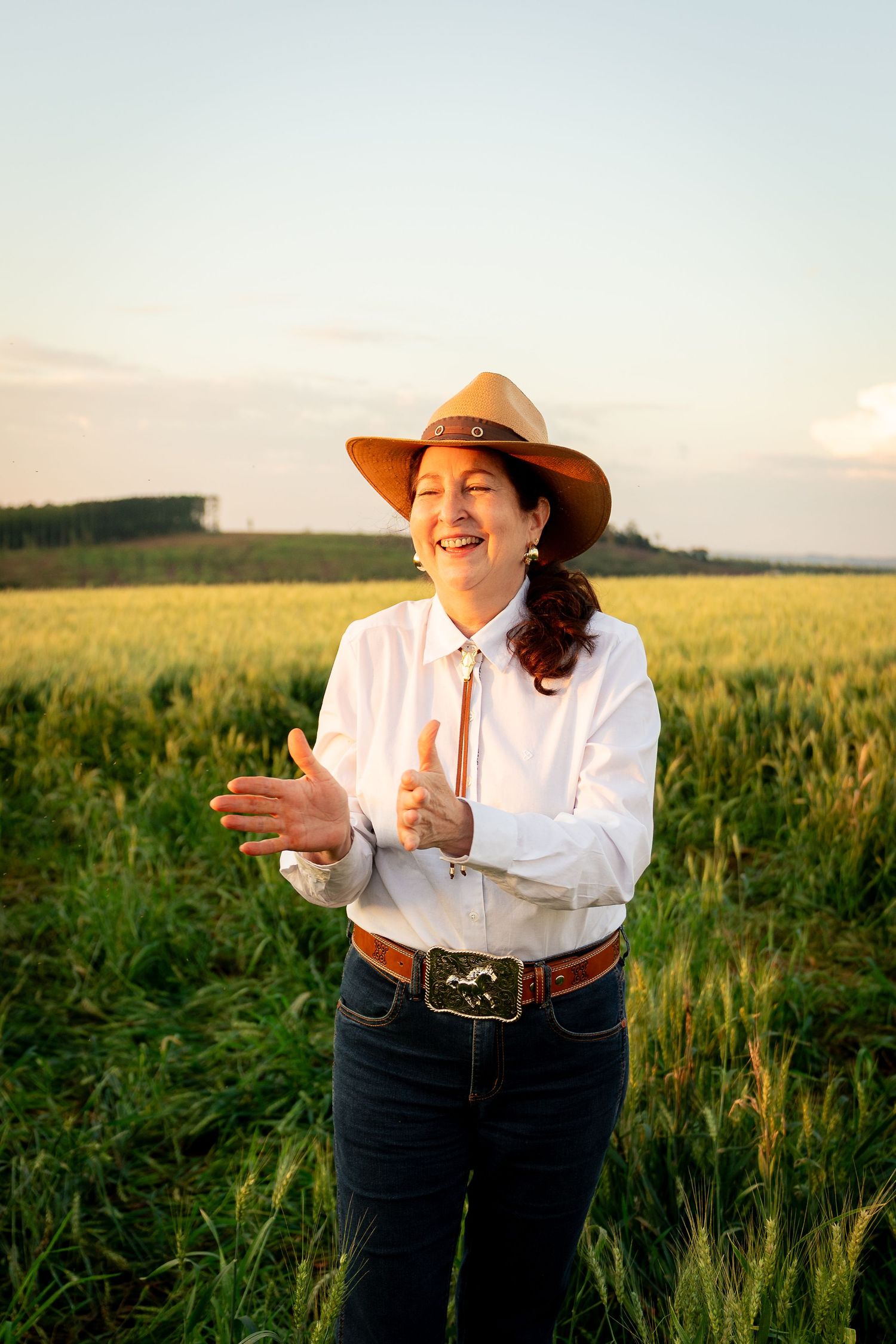 A smiling woman in a cowboy hat and white shirt claps her hands in a sunny green field at golden hour.