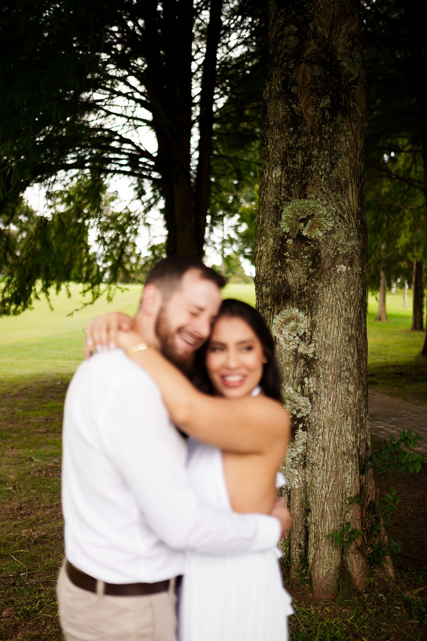 A smiling couple embraces near a mossy tree in a lush green park, wearing white and casual attire.