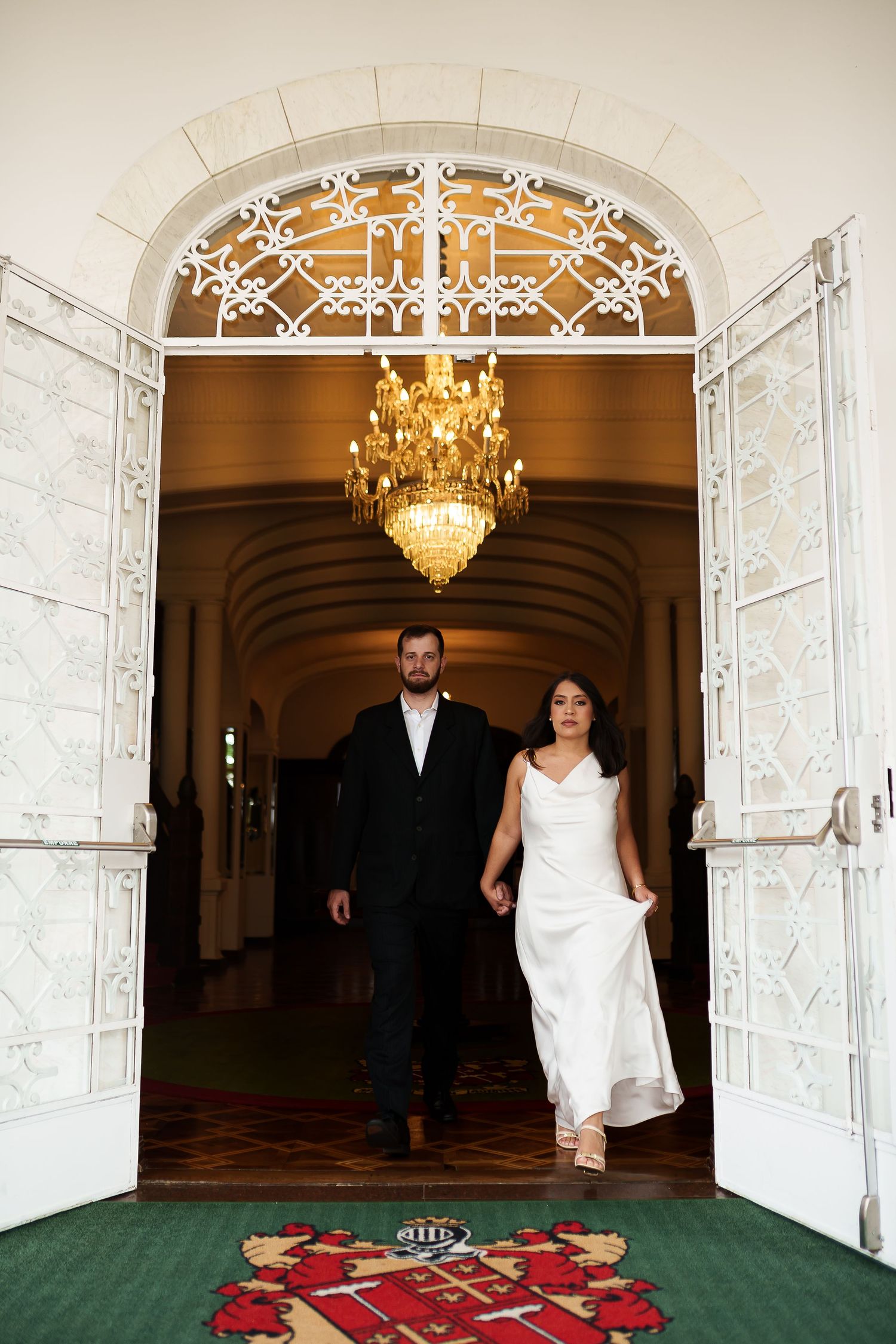 Bride and groom walking through elegant arched doorway with ornate white ironwork and golden chandelier.