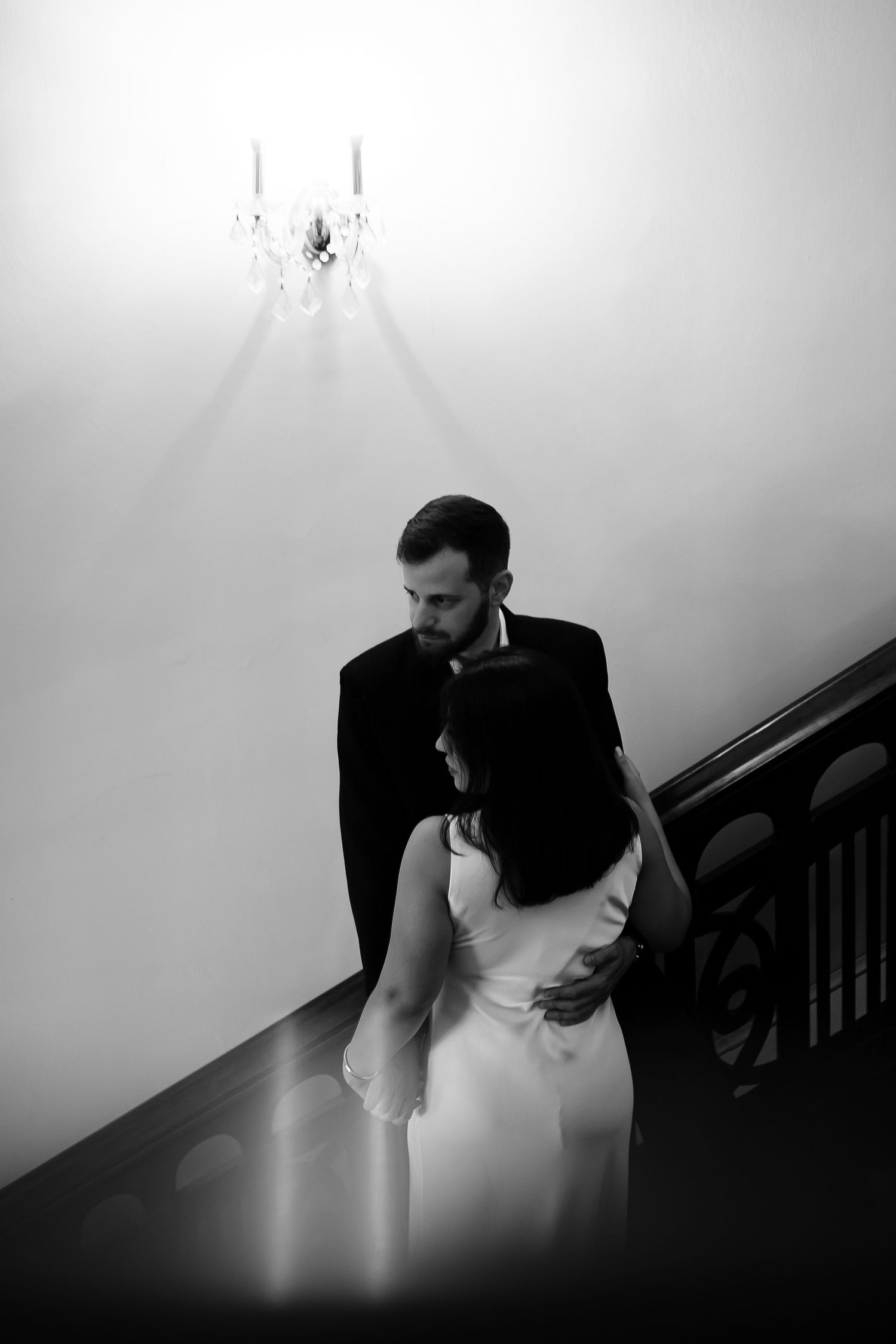 Black and white photo of an elegant couple embracing on a staircase beneath a crystal chandelier.