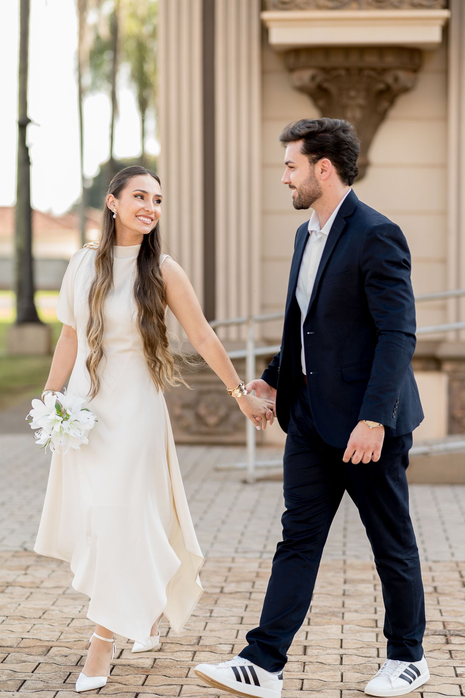 A smiling bride in white dress holds bouquet and groom's hand in navy suit walking outdoors.