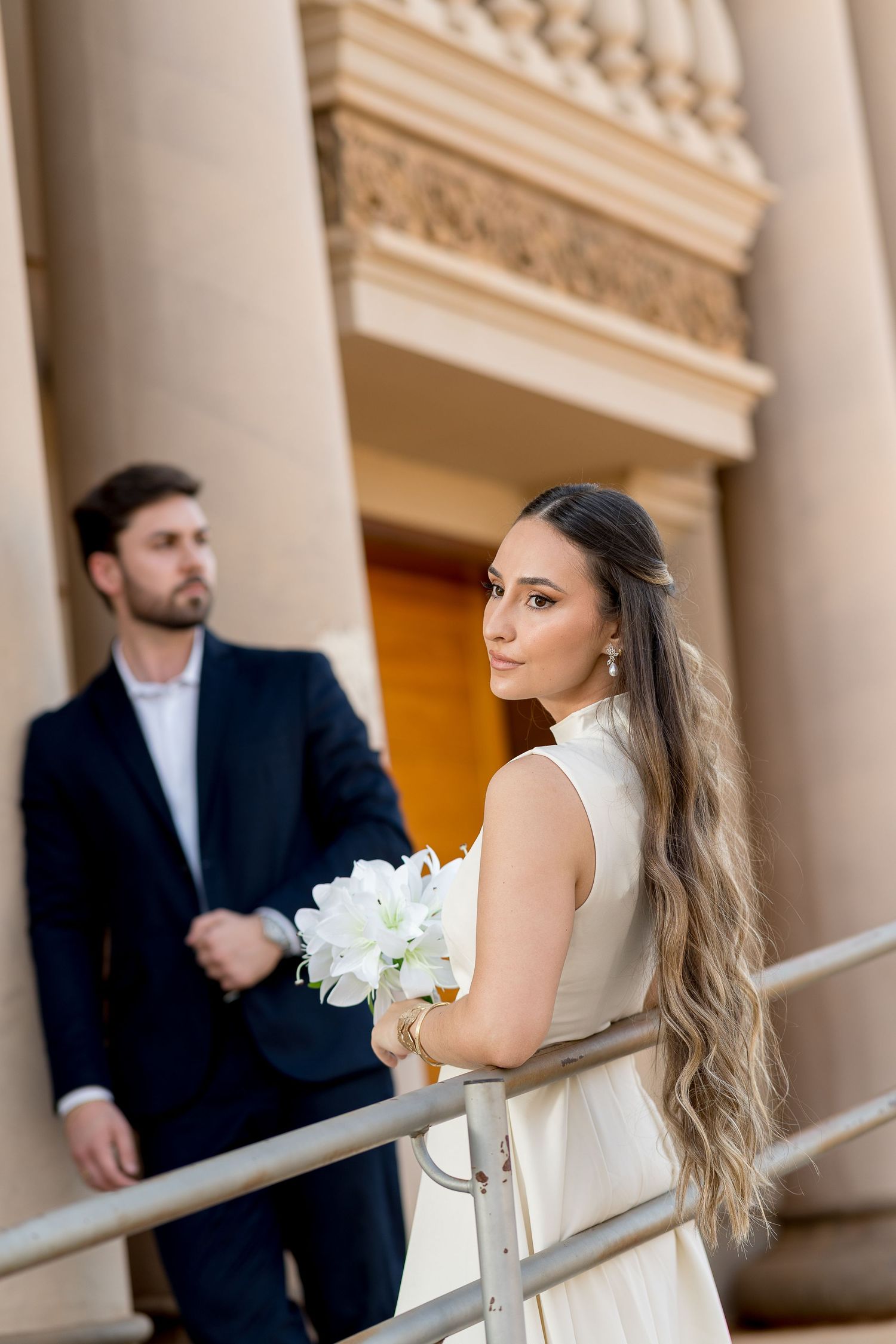 Elegant bride in white dress holding bouquet poses by railing with groom in dark suit near classical columns.