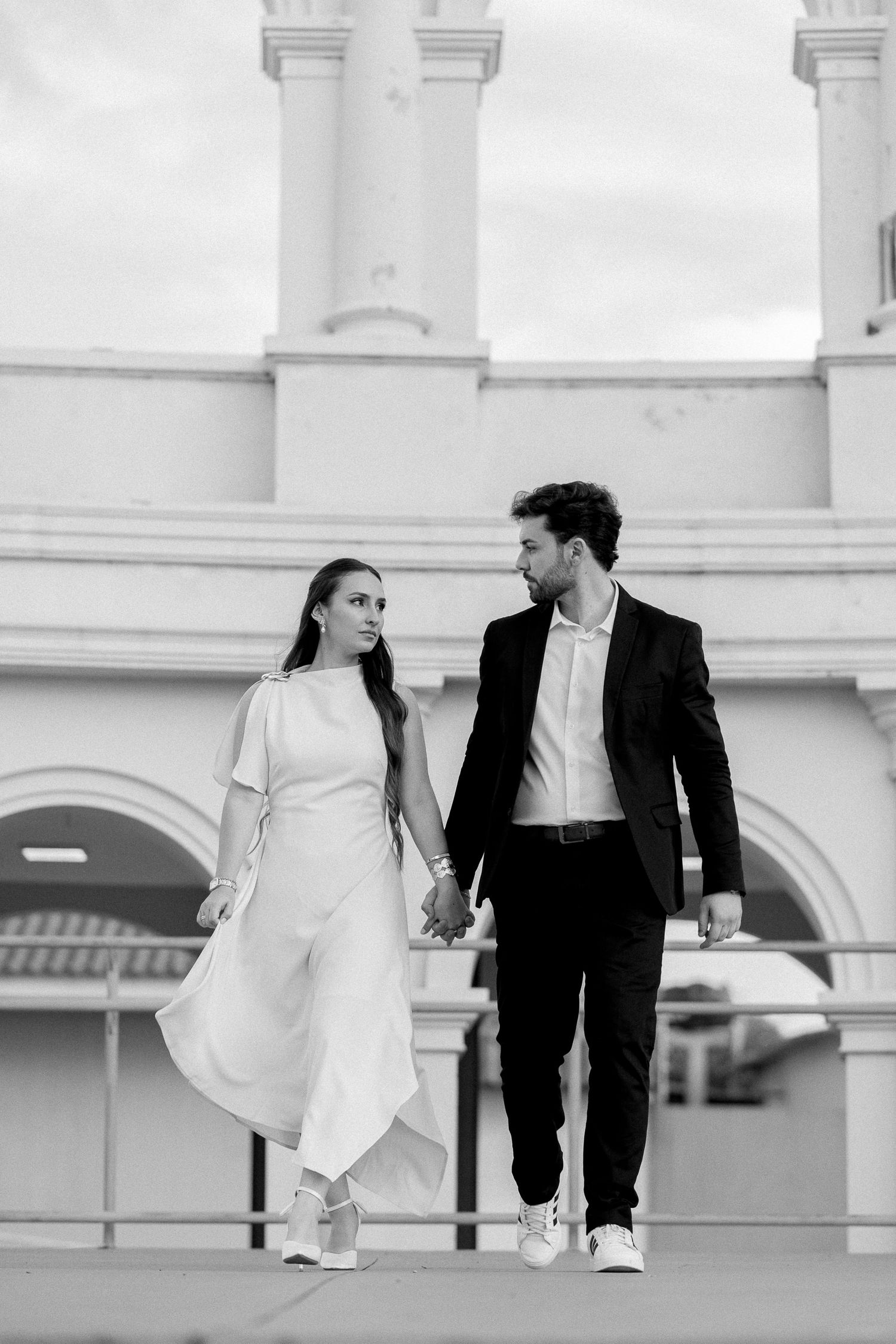 Black and white photo of an elegant couple holding hands, woman in white dress and man in black suit, walking together.