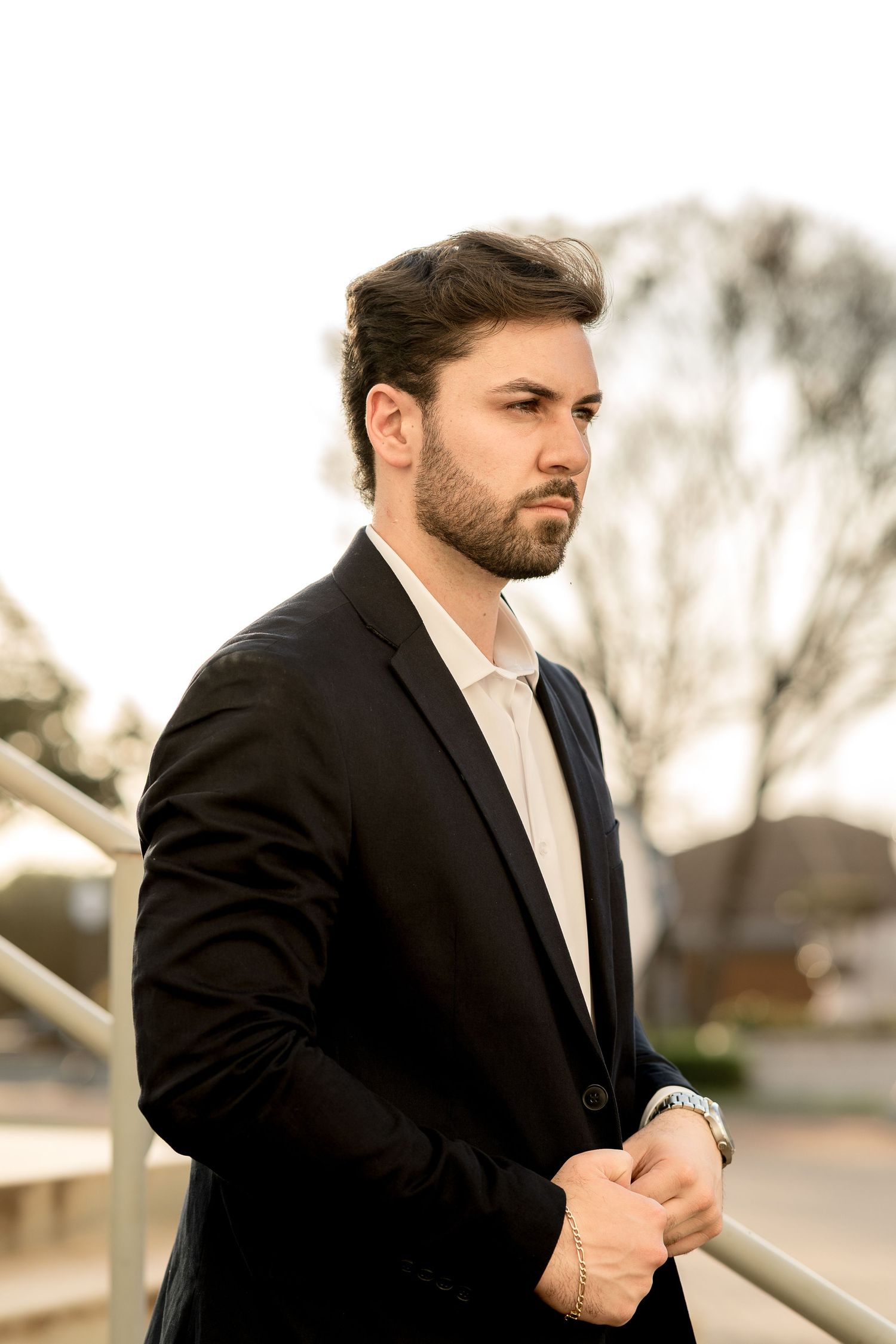 A well-dressed young man with a beard in a black blazer leans on a railing outdoors, looking thoughtfully to the side.