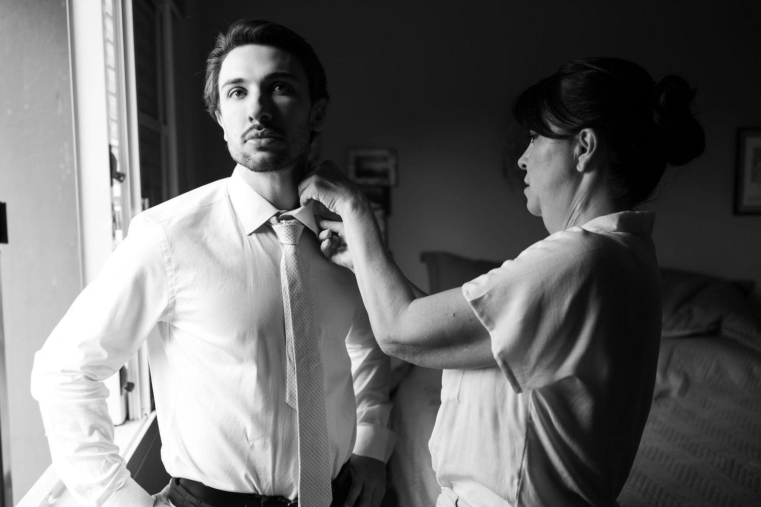 Black and white photo of a woman helping a groom adjust his tie while getting ready for a wedding.