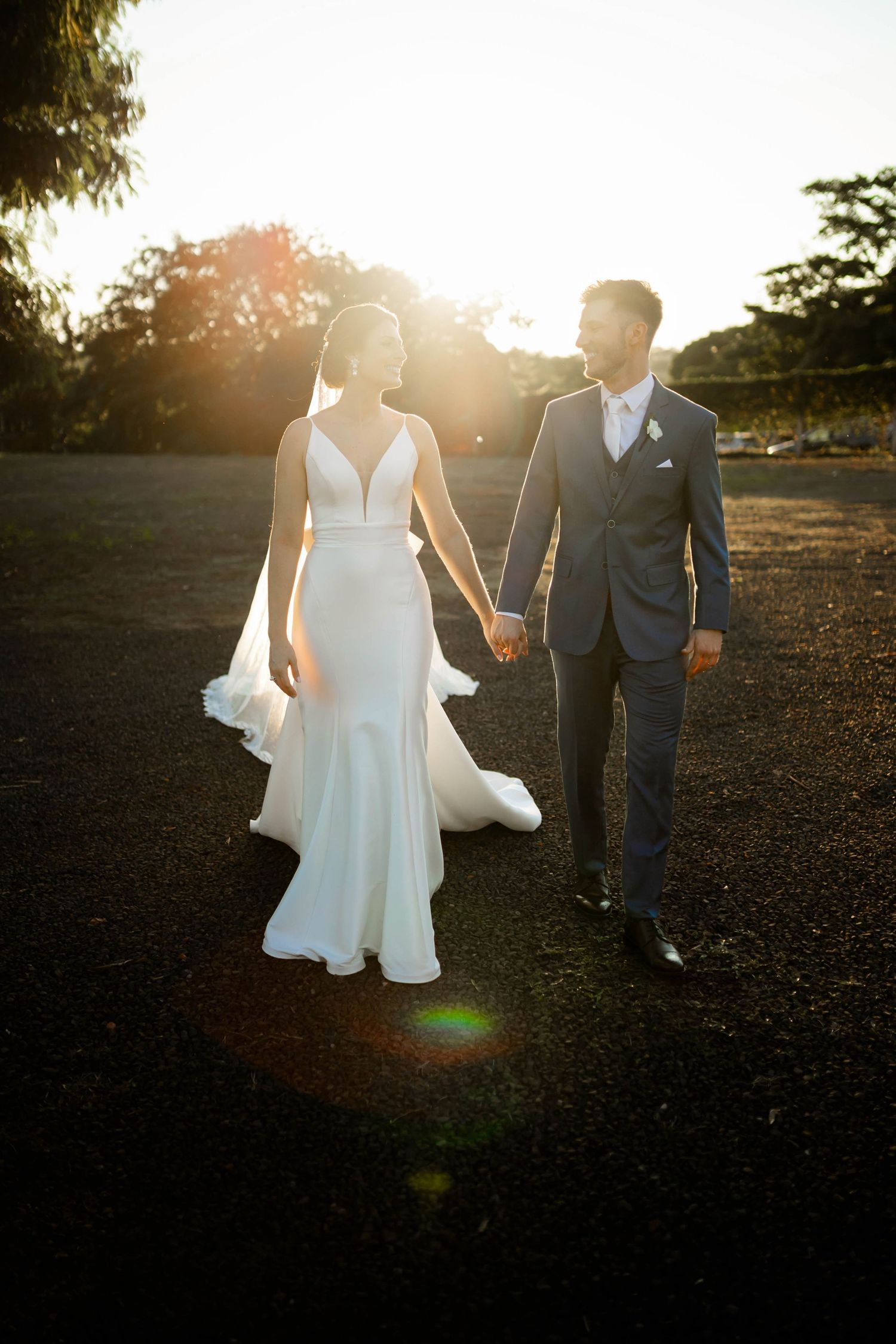 Bride and groom walk hand in hand at golden hour with sun flare behind them.