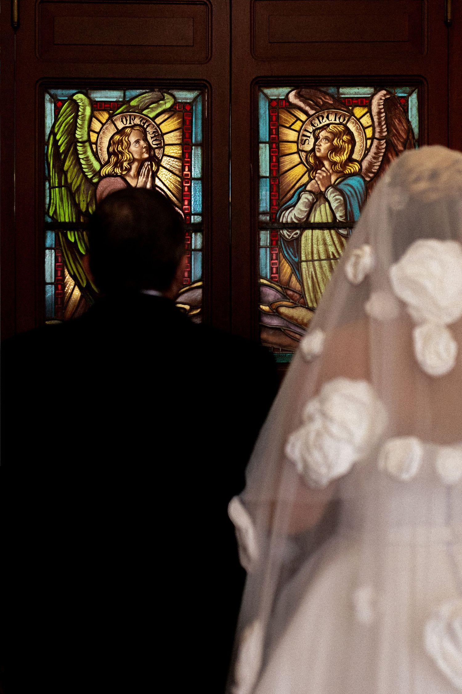 Bride and groom silhouetted before stunning angel stained glass church doors at wedding.