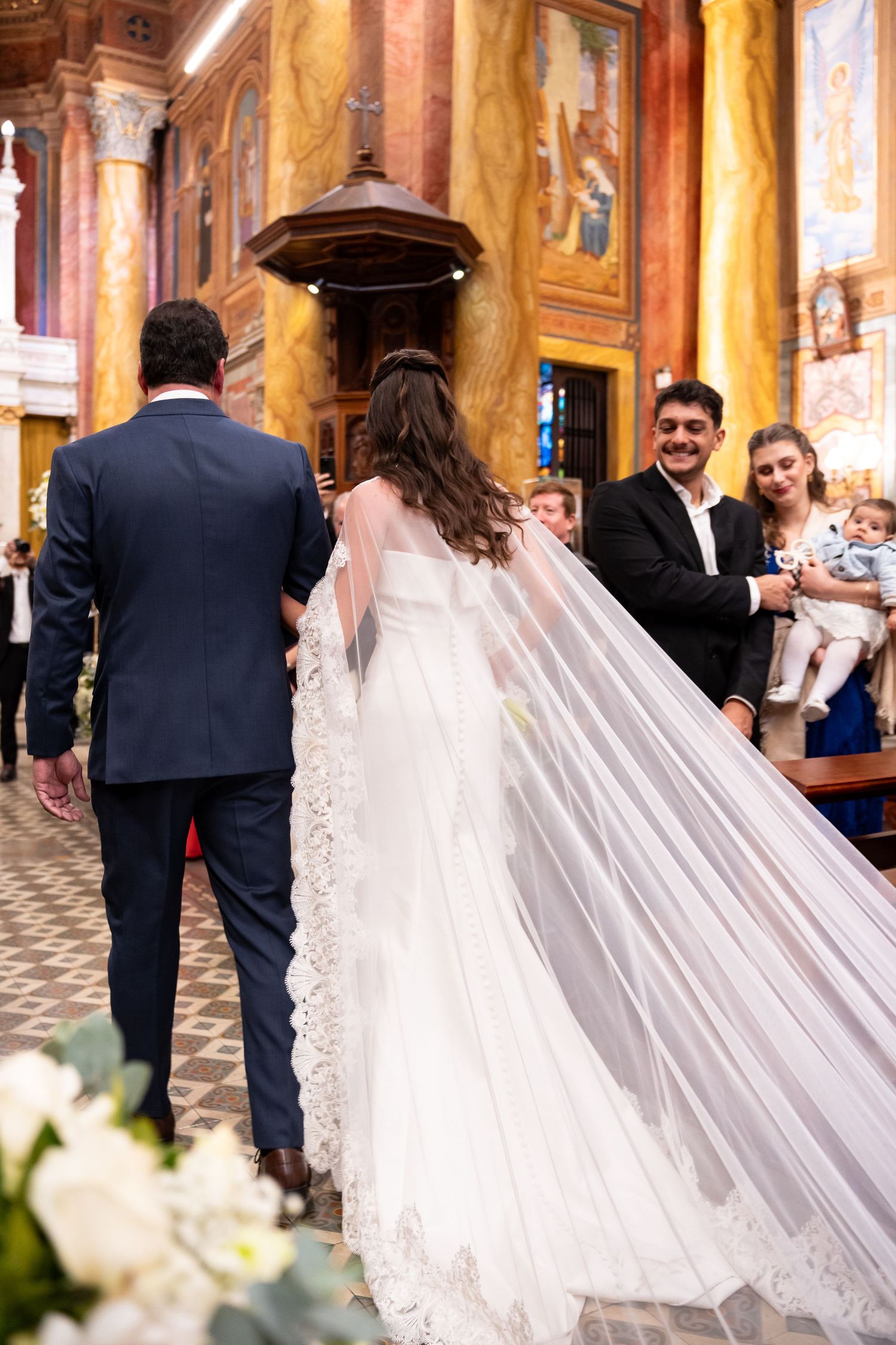 Bride and groom walking down church aisle with flowing lace veil, guests smiling in ornate golden chapel.