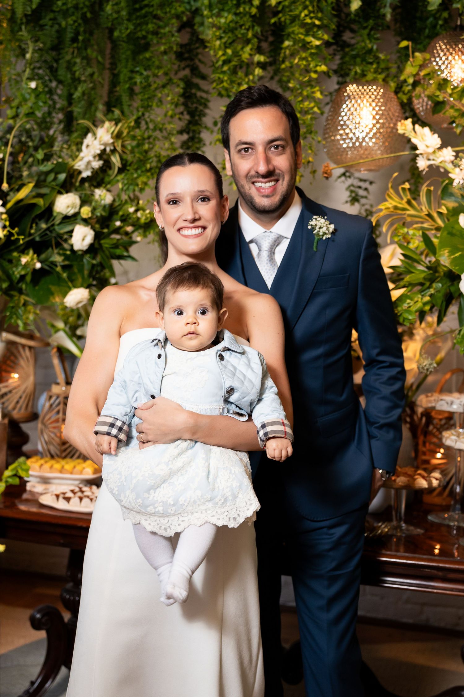 Smiling bride and groom in navy suit pose with baby at elegant wedding reception with greenery backdrop.
