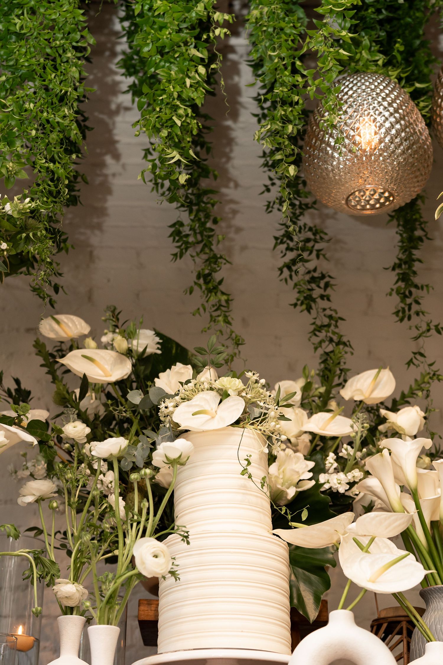 Elegant white floral arrangement with calla lilies, ranunculus, and hanging greenery surrounding a striped white vase.