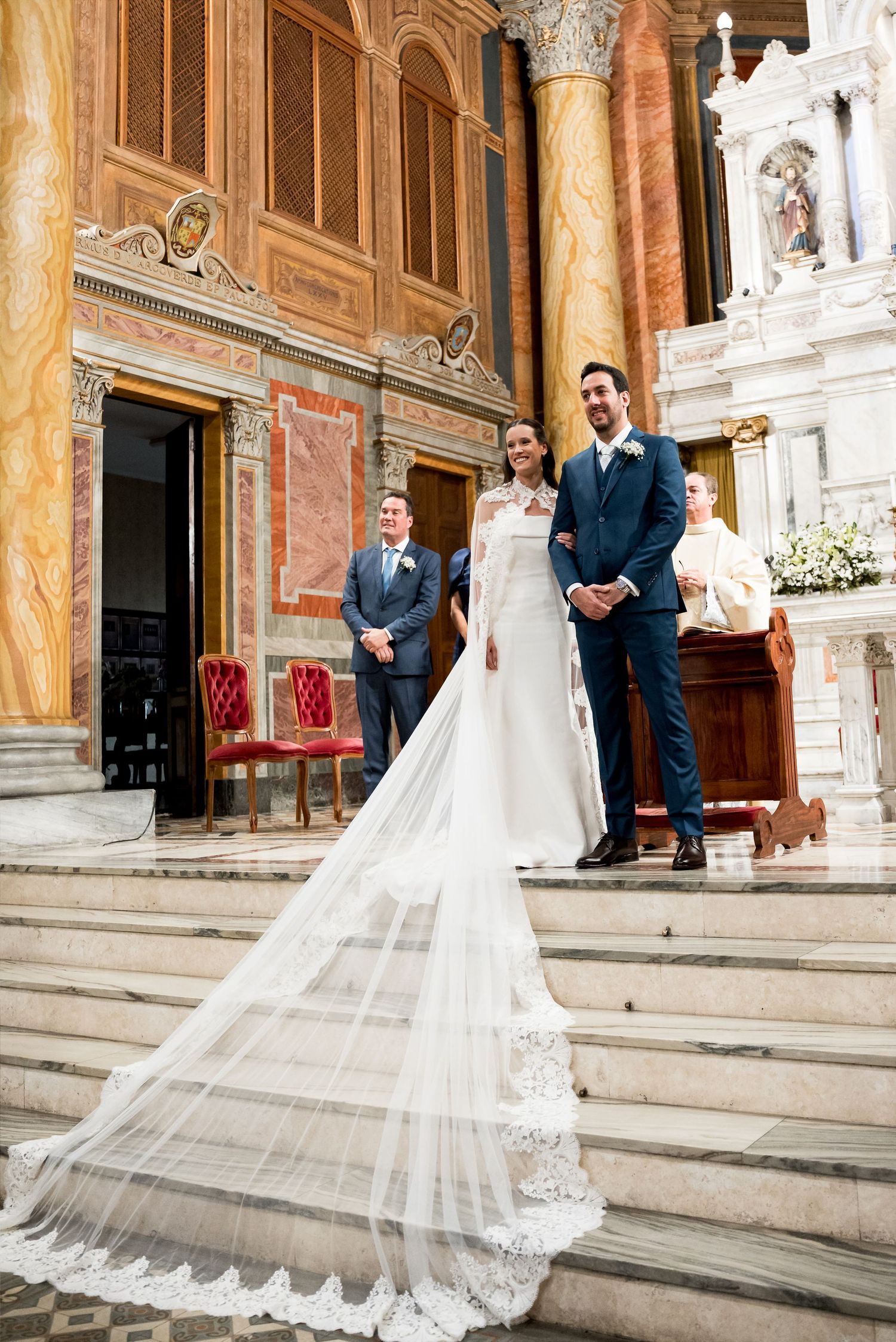 Bride in elegant gown with long lace veil stands with groom in navy suit on church steps during wedding ceremony.