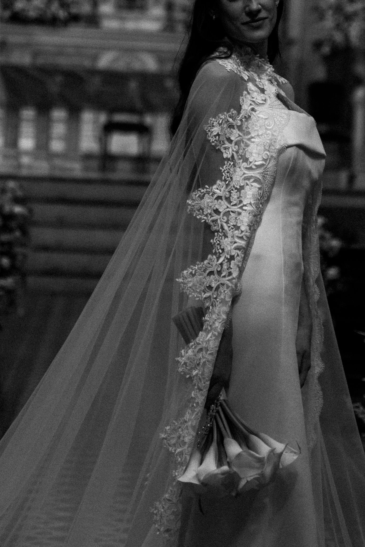 Black and white photo of a bride in an elegant lace wedding gown with a flowing veil cape holding a bouquet.