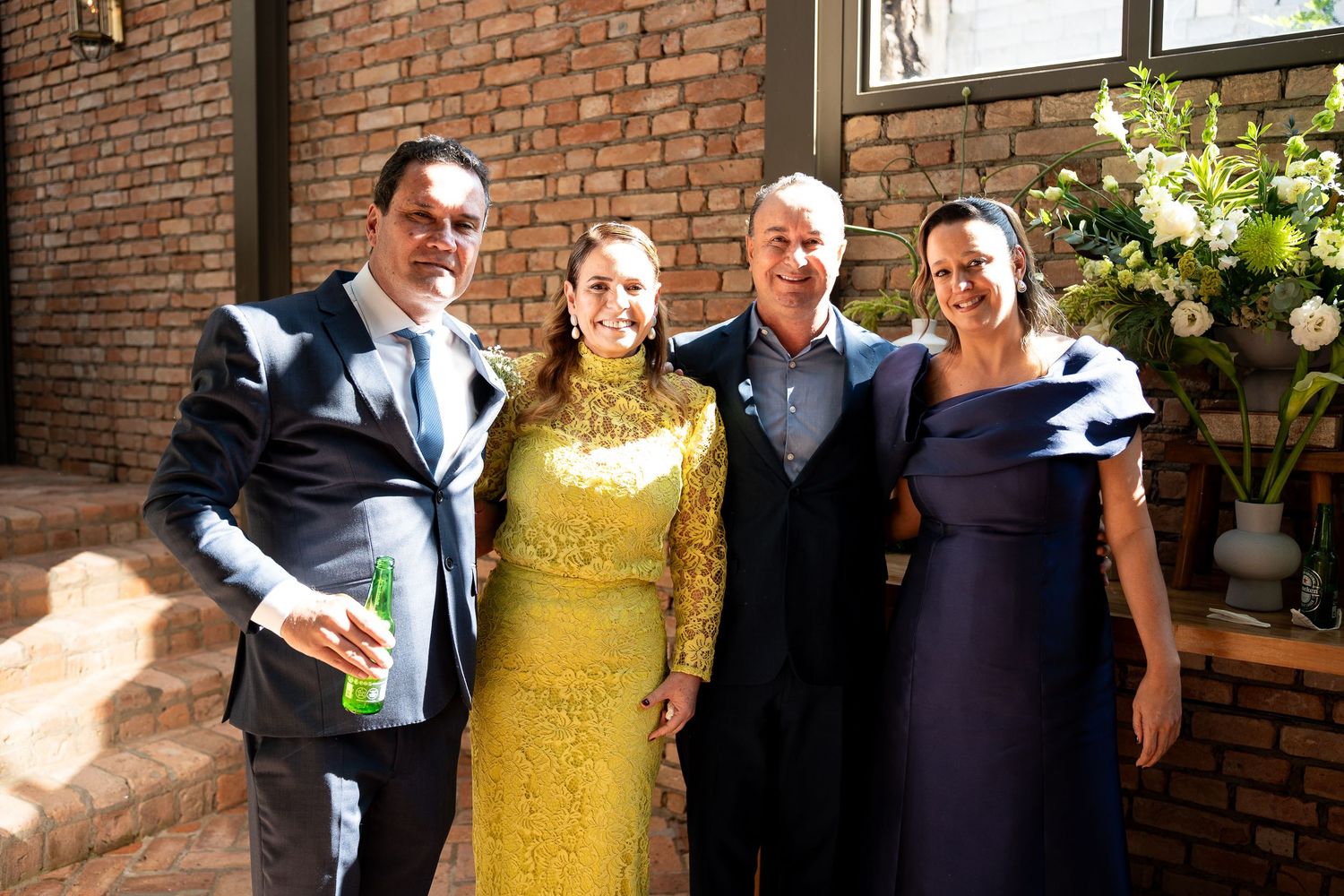 Four elegantly dressed guests posing together at a formal outdoor event near a brick wall with floral arrangements.