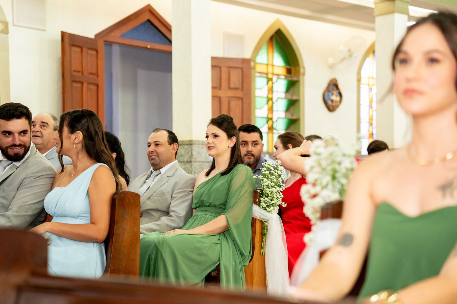 Guests seated in church pews during a wedding ceremony, wearing elegant attire in green, blue, and red.