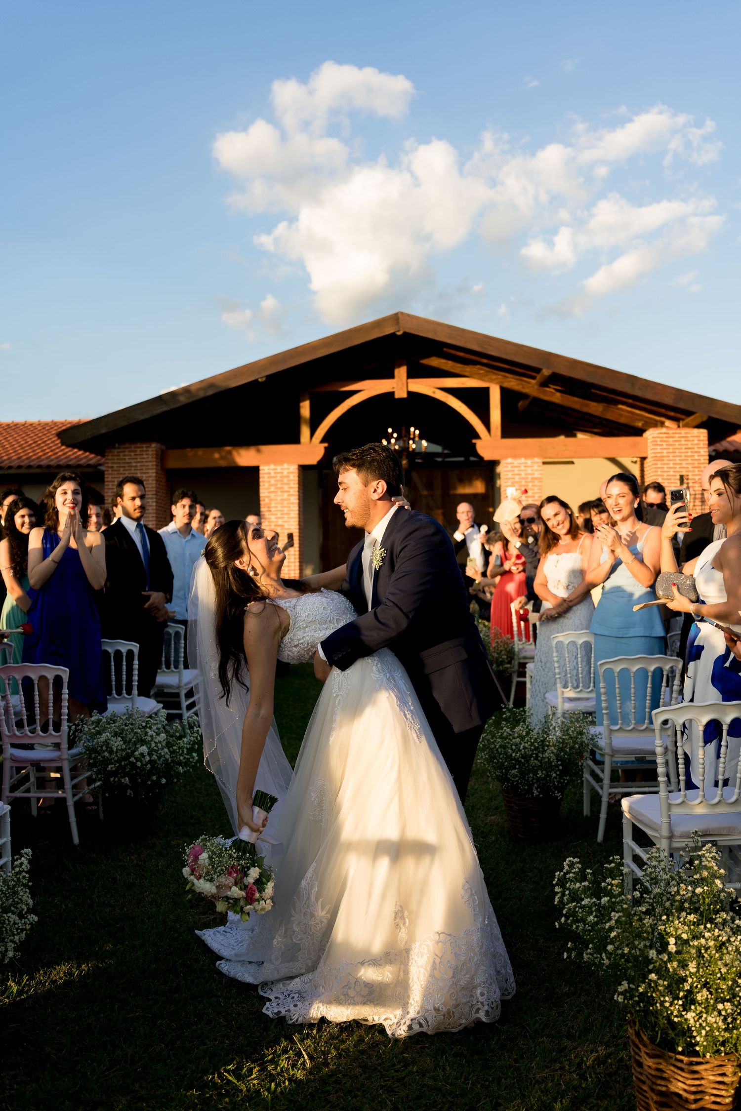 Groom lifts bride in white gown during outdoor wedding ceremony while guests applaud beside white chairs.