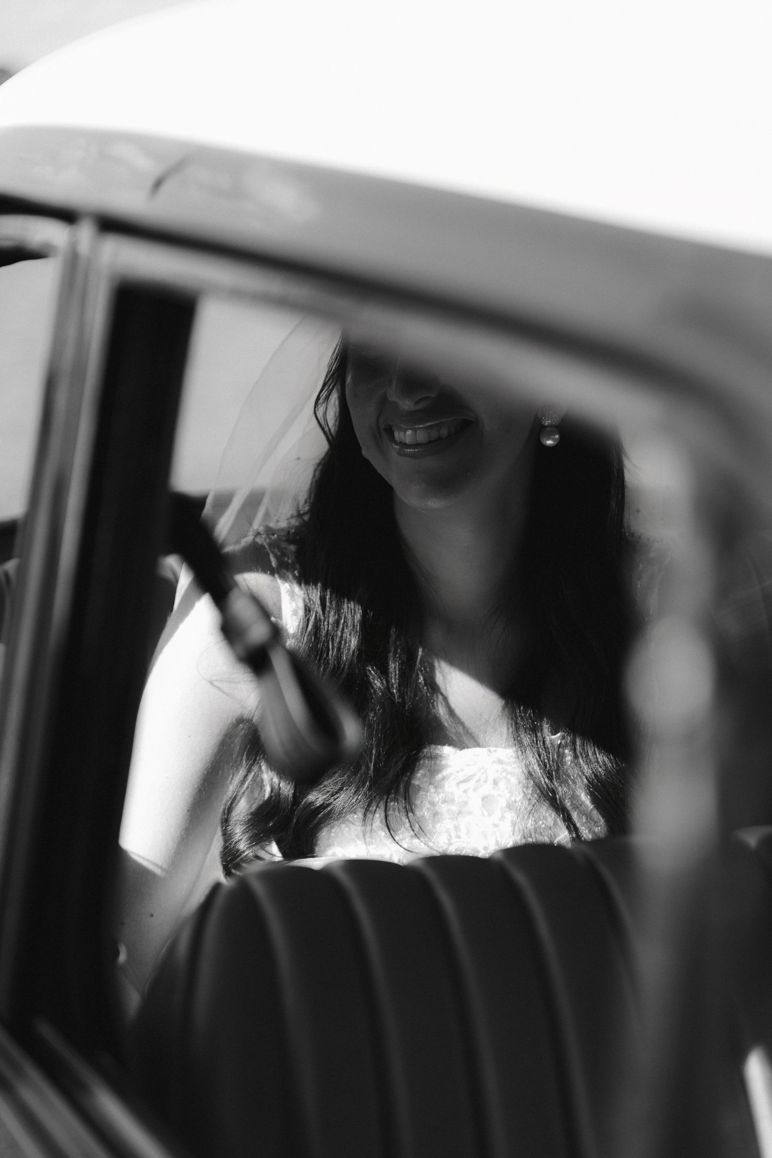 Black and white photo of a smiling bride in a wedding dress sitting in a vintage car, looking at her phone.