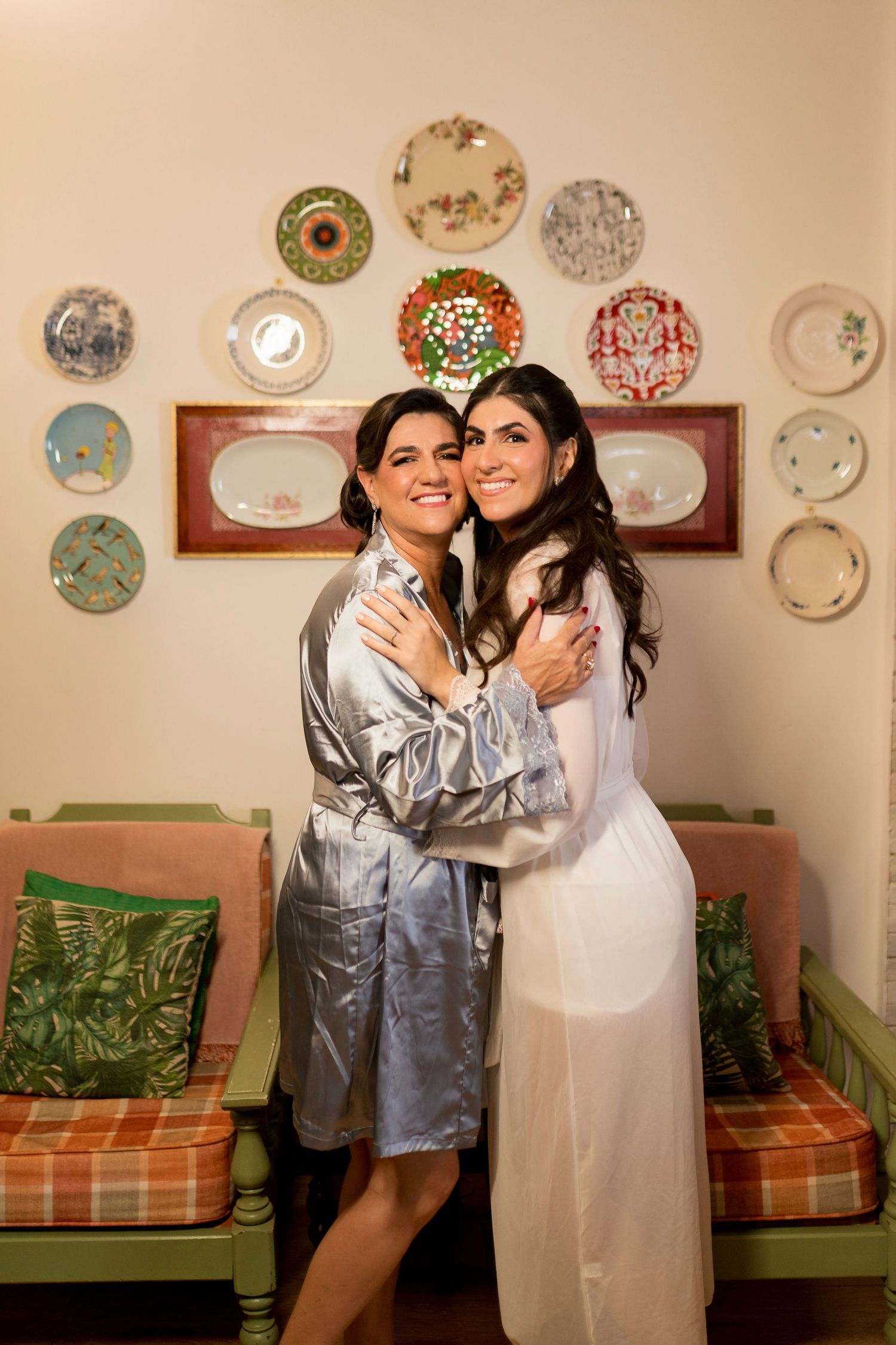 Two women in robes embrace and smile in a decorated room with colorful plates on the wall behind them.